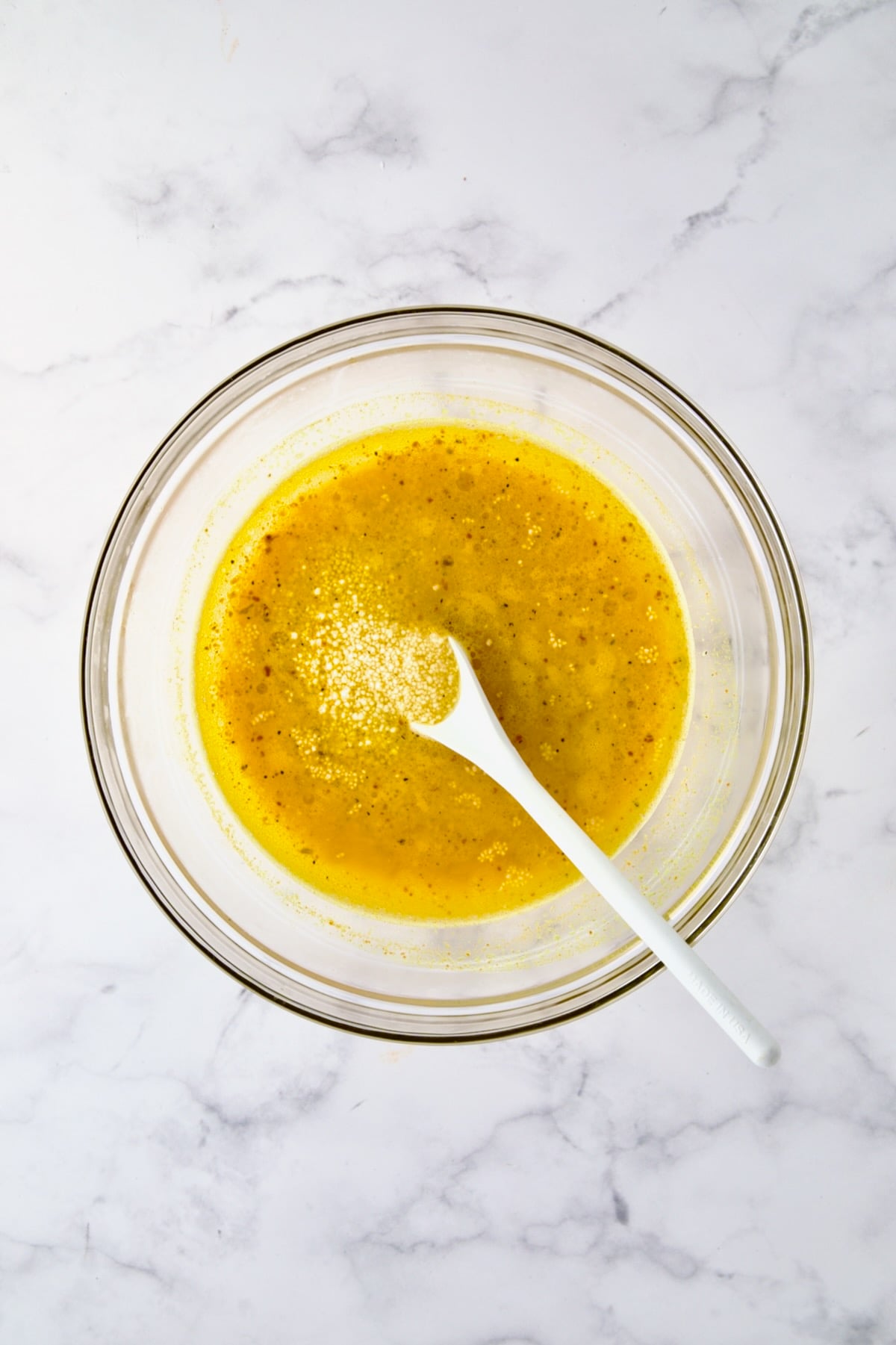 Uncooked couscous added to water and seasonings in glass bowl being stirred with white mixing spoon.