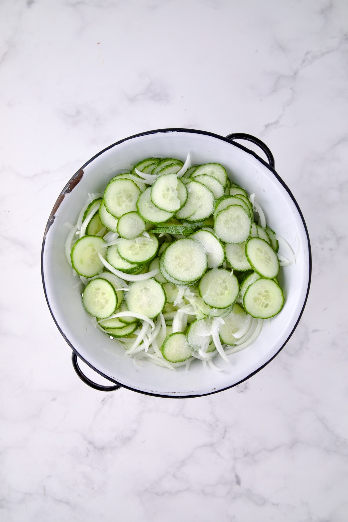 Cucumbers and onions in white distressed colander sprinkled with salt.