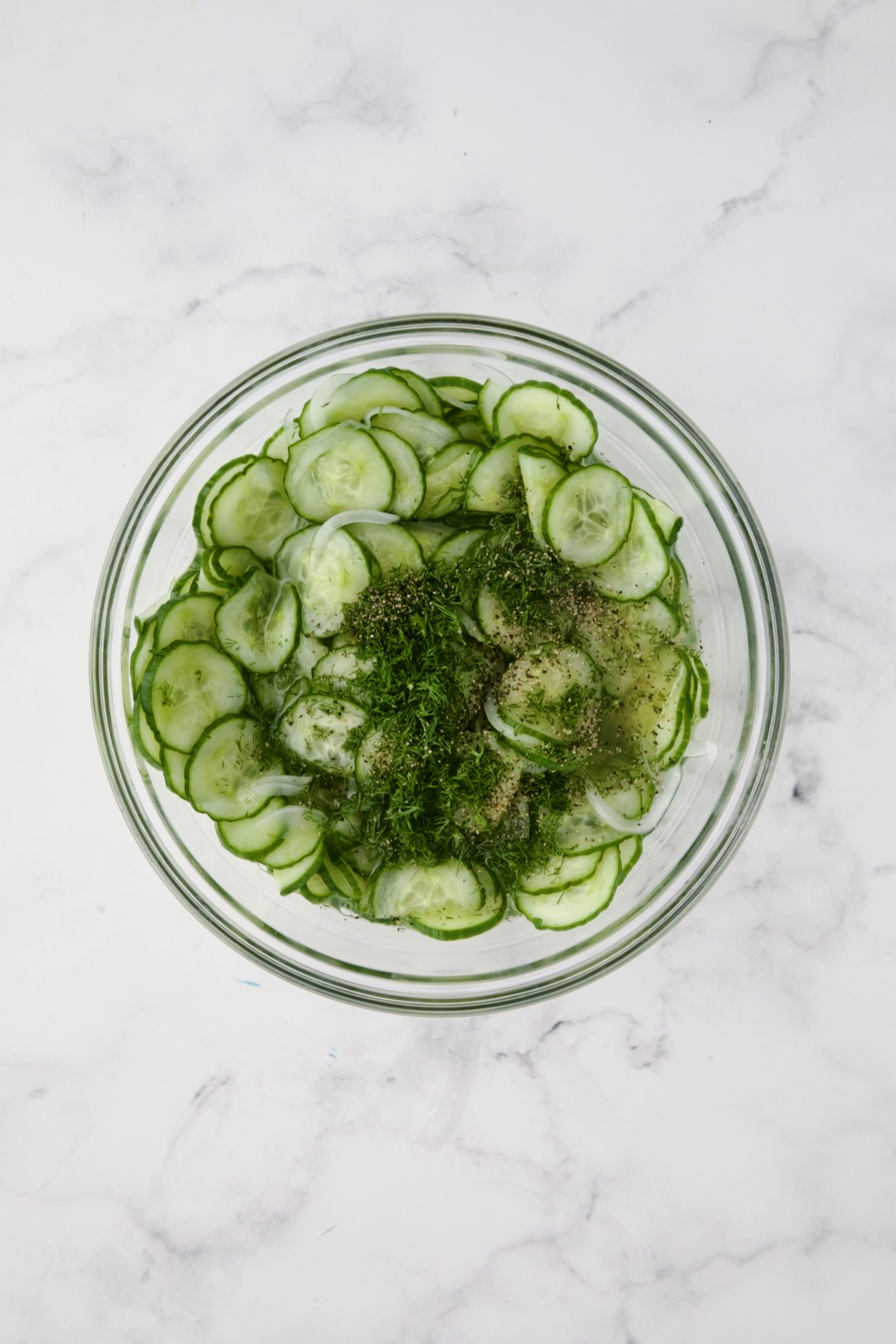 Cucumbers, onions, dressing and dill in glass bowl.