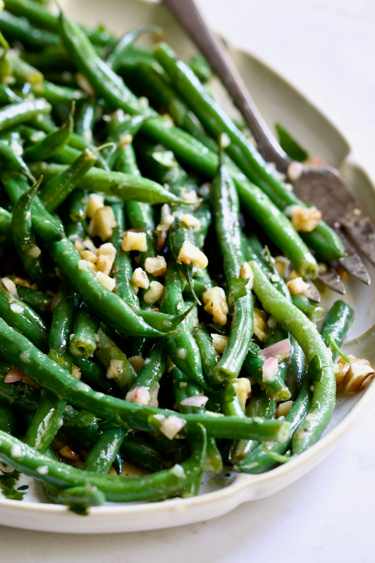 Green Bean Salad with Mustard Vinaigrette on white oval platter with serving utensils garnished with walnuts.