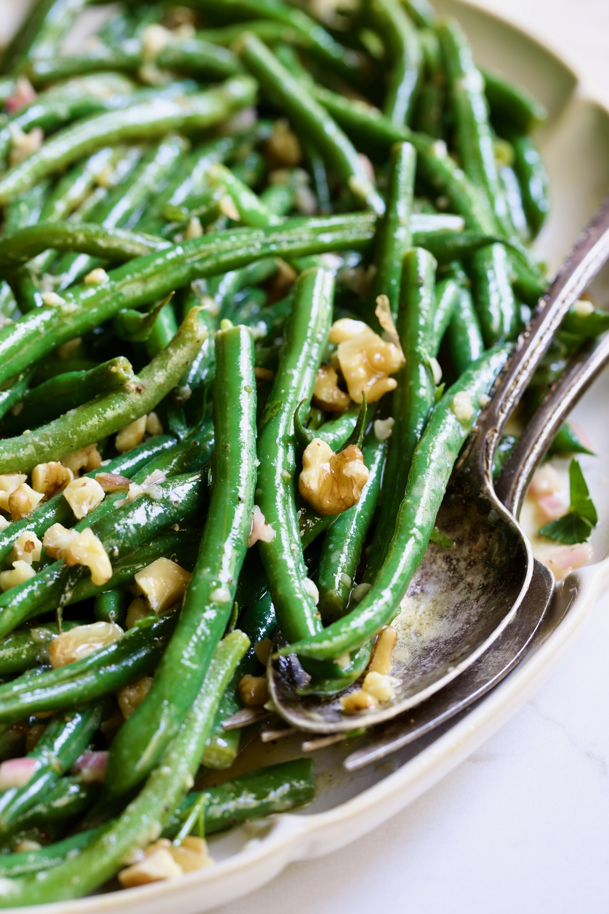 Green Bean Salad with Mustard Vinaigrette on white oval platter with serving utensils garnished with walnuts.