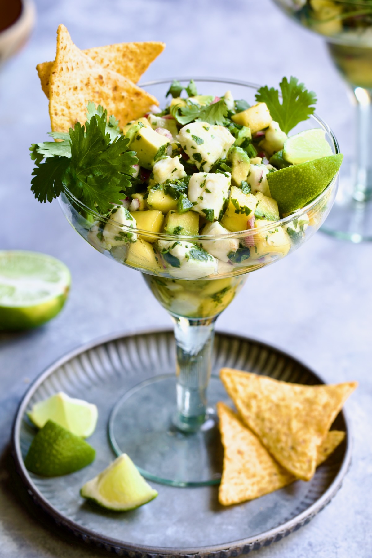 Halibut Ceviche with mango, avocado, lime wedges, cilantro sprigs and tortilla chips in margarita glass.