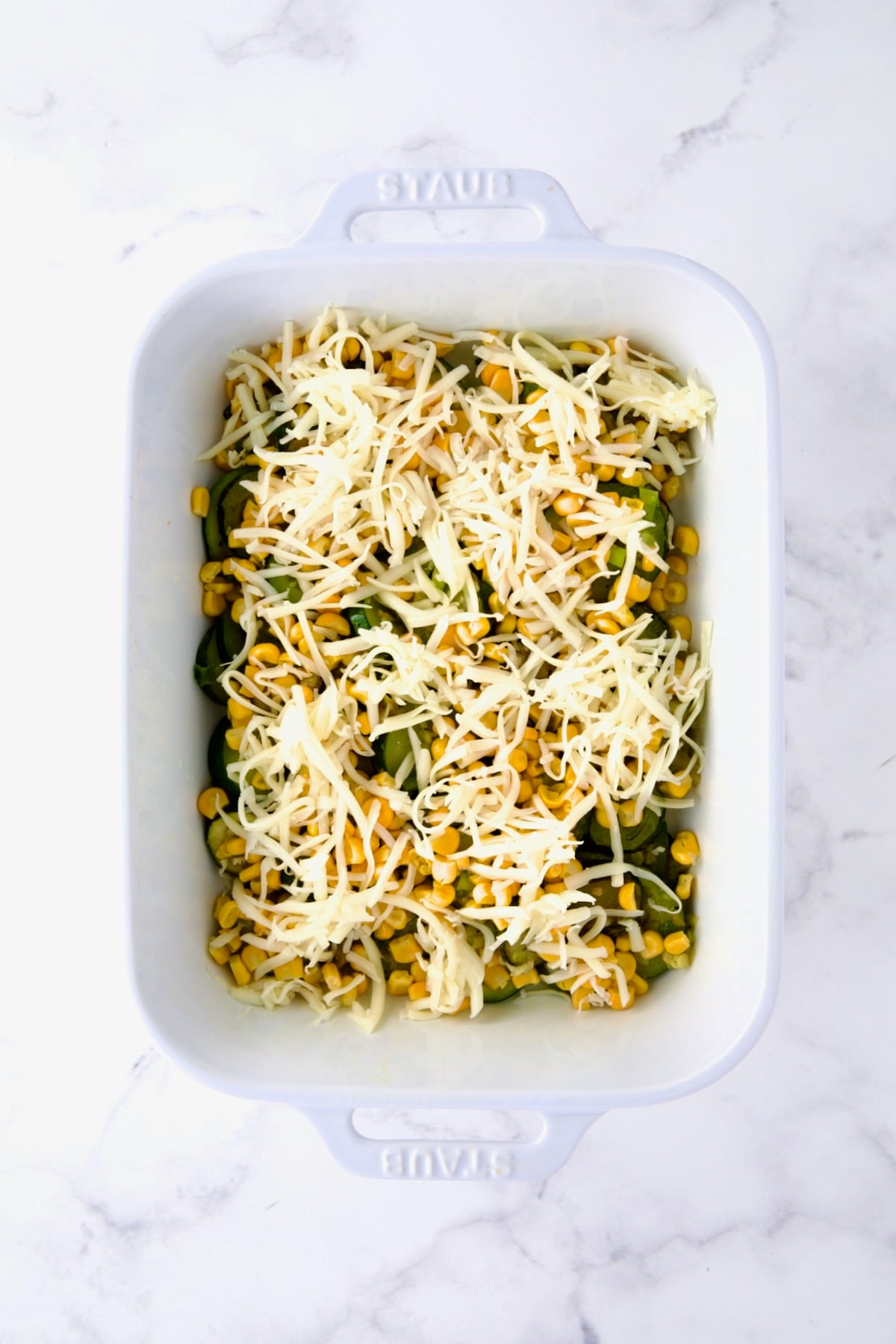 Cheese sprinkled over zucchini and corn in white rectangular baking dish.