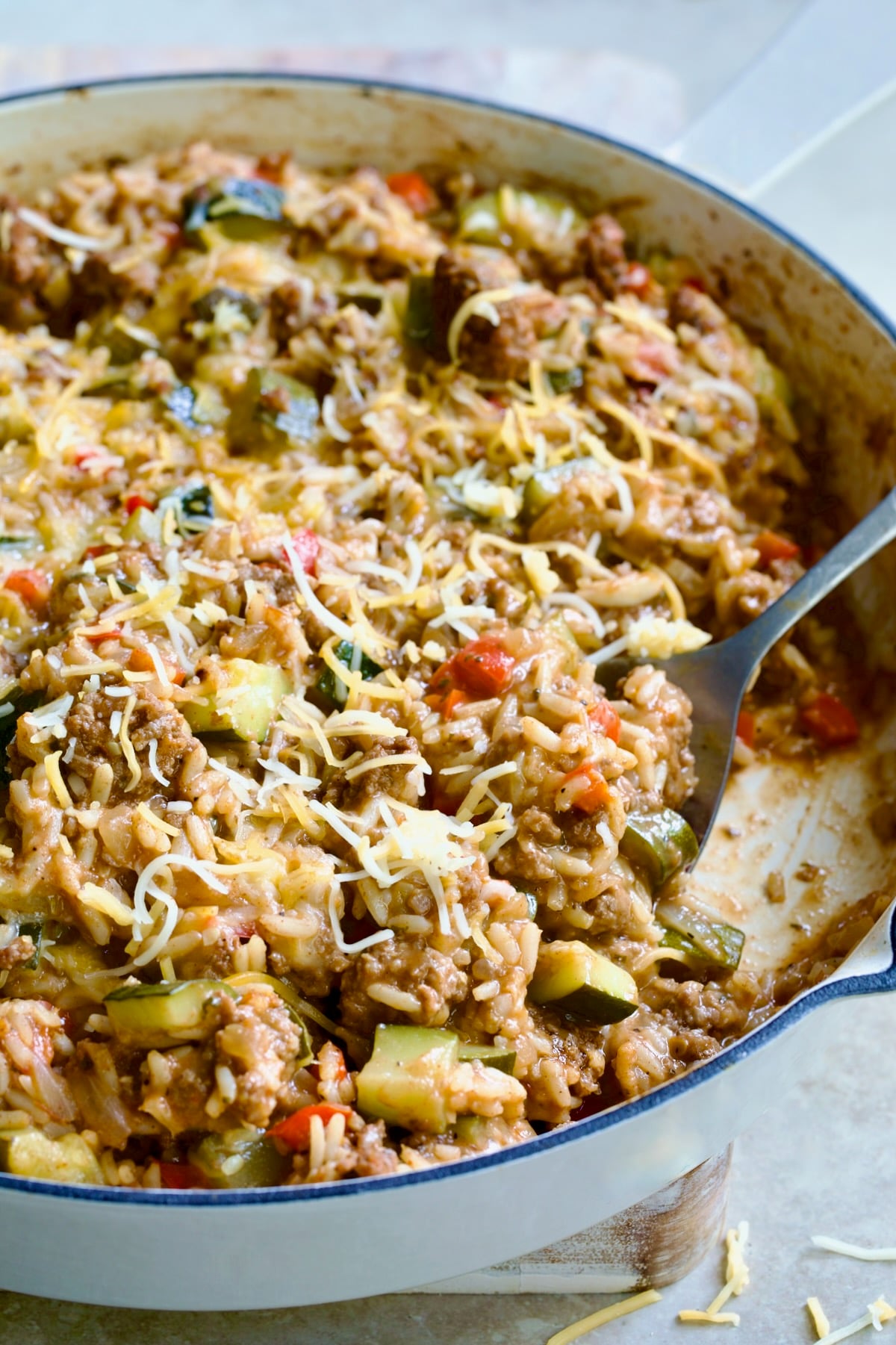Beef Zucchini Rice Skillet in white enamel cast iron skillet on white piece of wood with serving spoon scooping some up.
