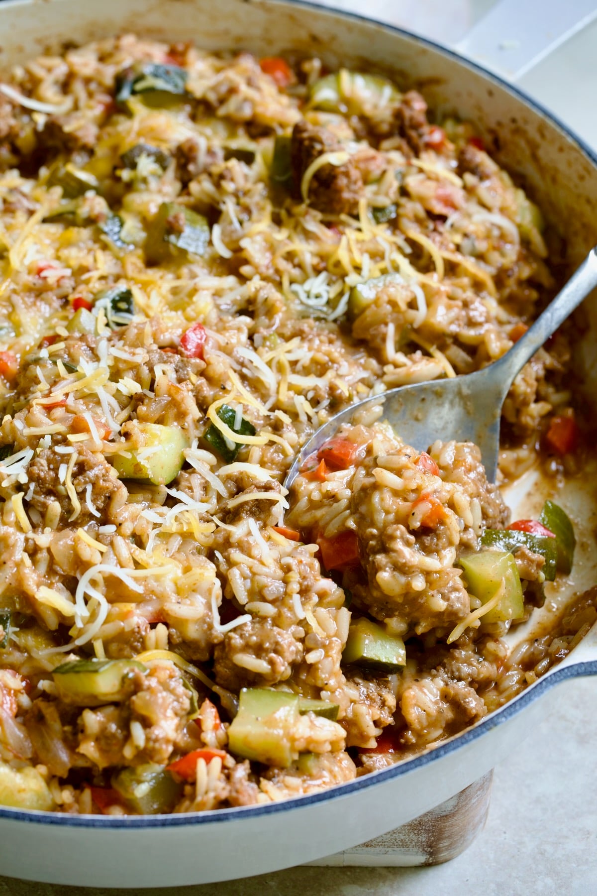 Beef Zucchini Rice Skillet in white enamel cast iron skillet on white piece of wood with serving spoon scooping some up.