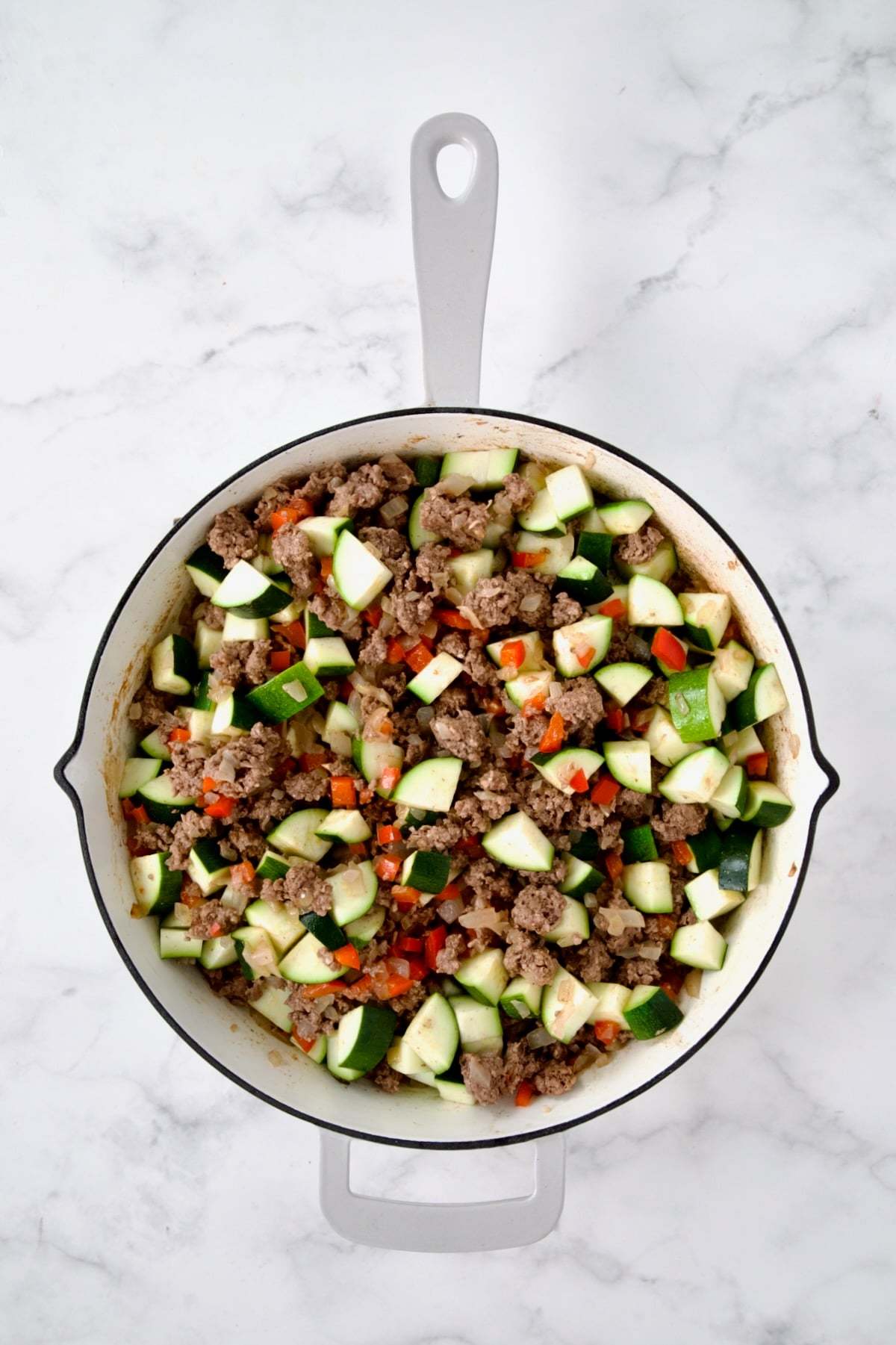 Cooked ground beef, red bell pepper and onions with zucchini added in white enamel cast iron skillet.