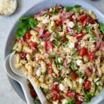 Caprese Pasta Salad in blue bowl with serving utensils with small bowls of Parmesan cheese, crushed red pepper flakes and fresh basil near it.
