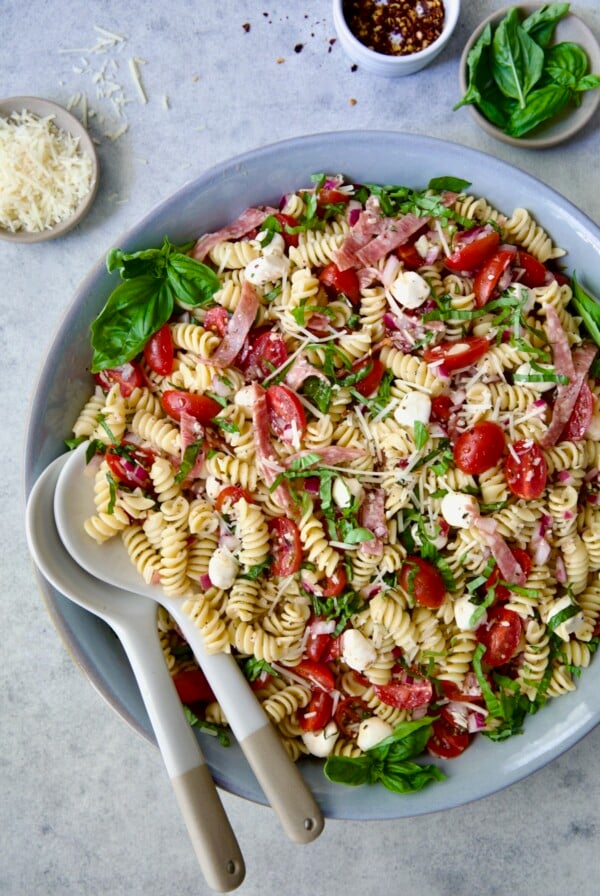 Caprese Pasta Salad in blue bowl with serving utensils with small bowls of Parmesan cheese, crushed red pepper flakes and fresh basil near it.