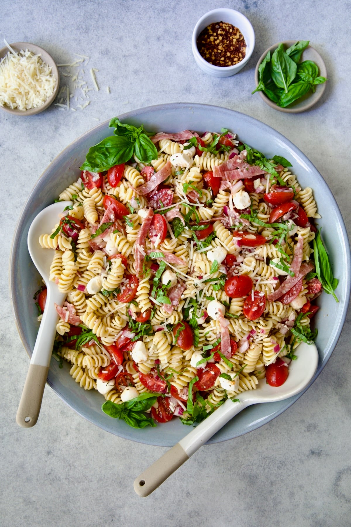 Caprese Pasta Salad in blue bowl with serving utensils with small bowls of Parmesan cheese, crushed red pepper flakes and fresh basil near it.