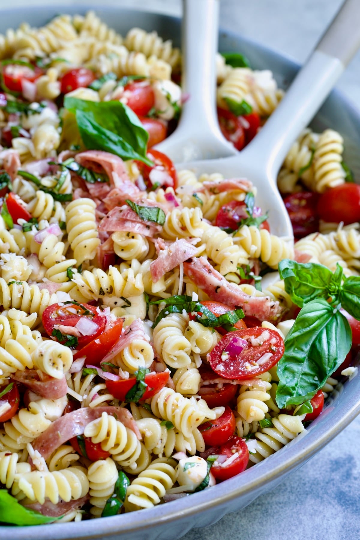Caprese Pasta Salad in blue bowl being stirred with serving utensils.