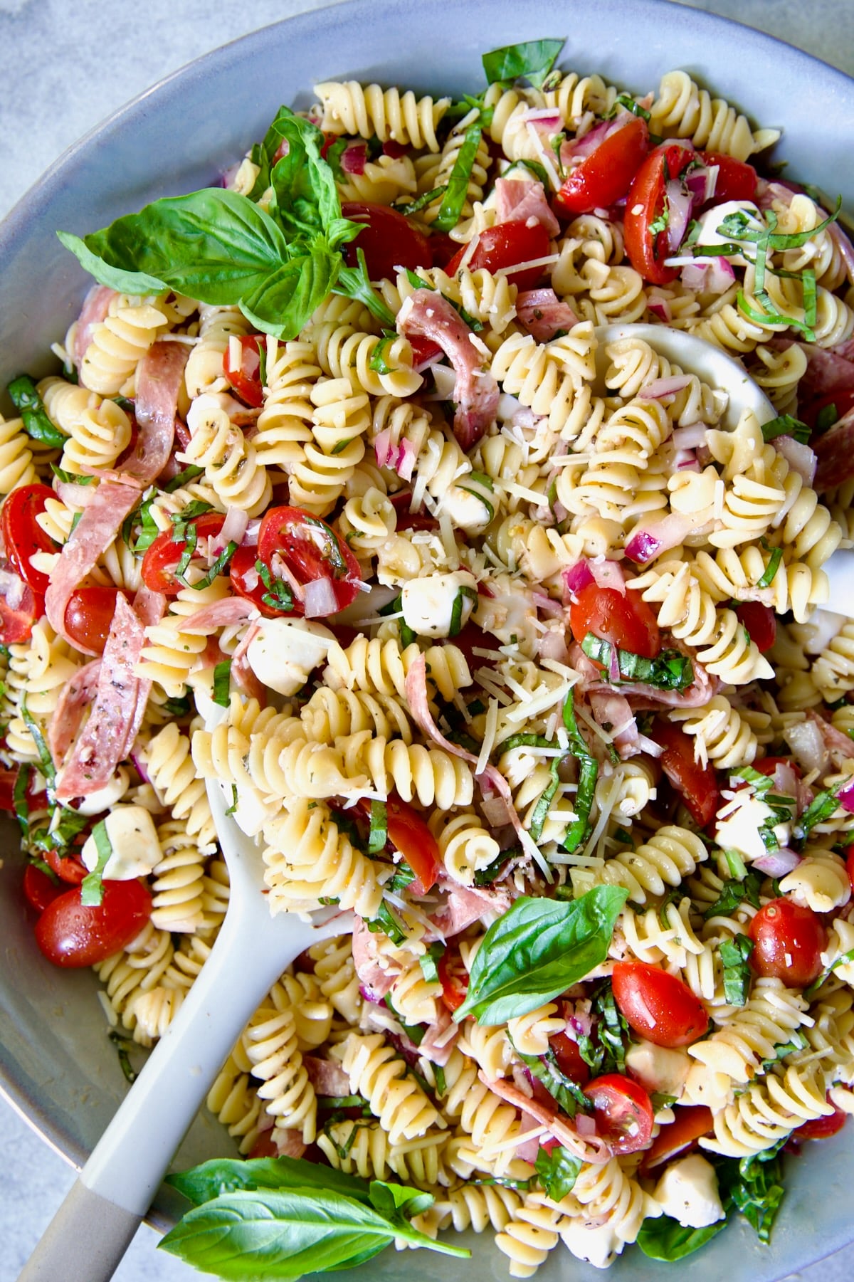 Caprese Pasta Salad in blue bowl being stirred with serving utensils.