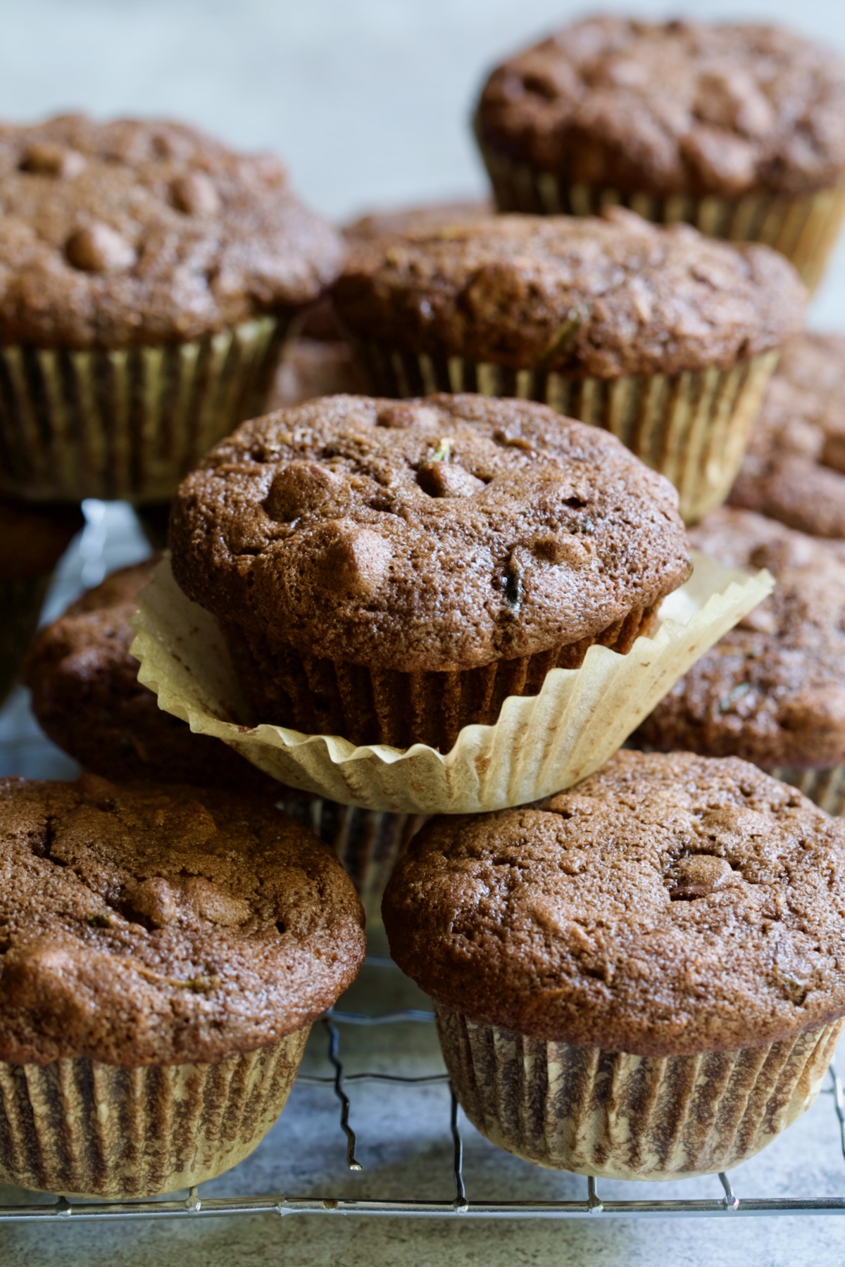 Pile of Chocolate and Zucchini Muffins on cooling rack with paper liner pulled away from one of them.