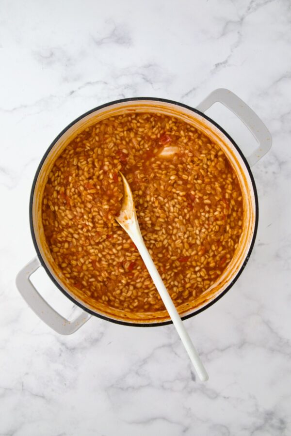 Cooked Barley Vegetable Risotto in white enamel cast iron skillet being stirred with white mixing spoon.