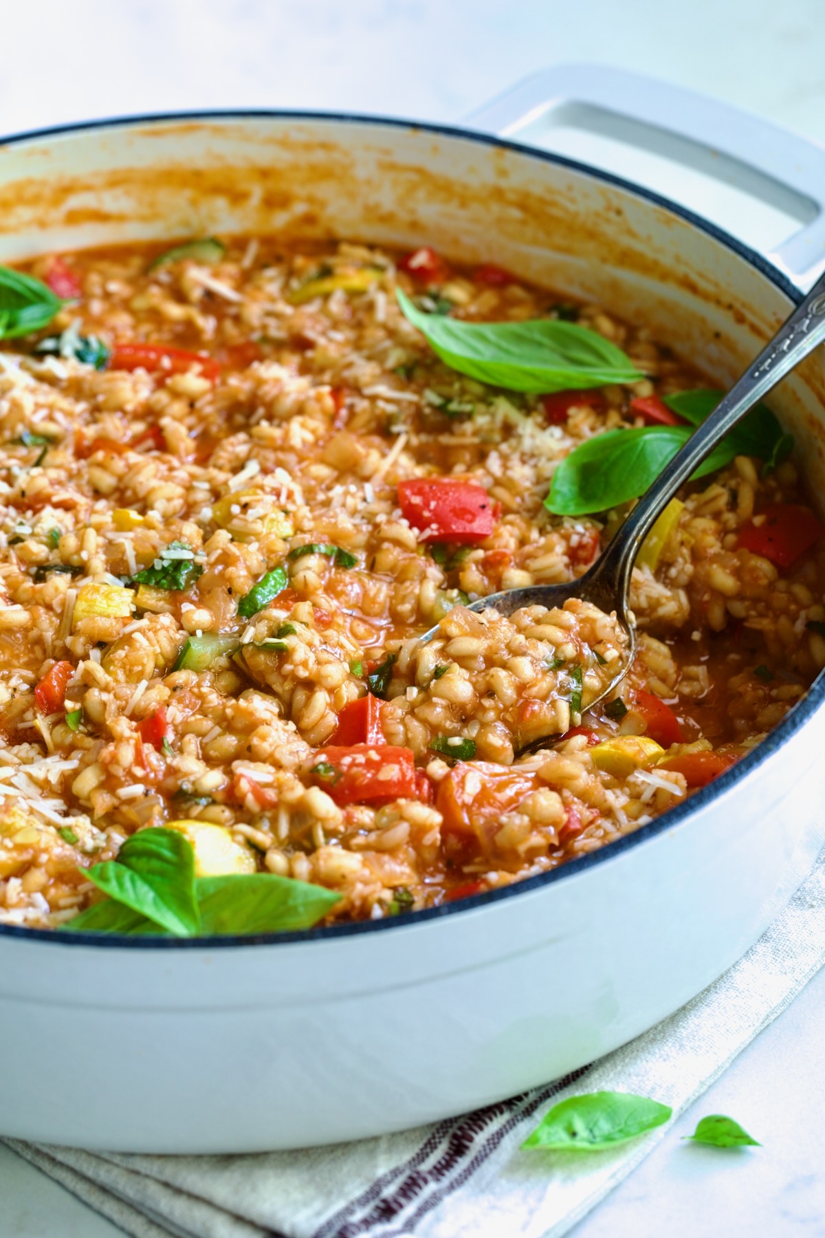 Barley Vegetable Risotto in white enameled cast iron pan garnished with fresh basil with serving spoon scooping some up.