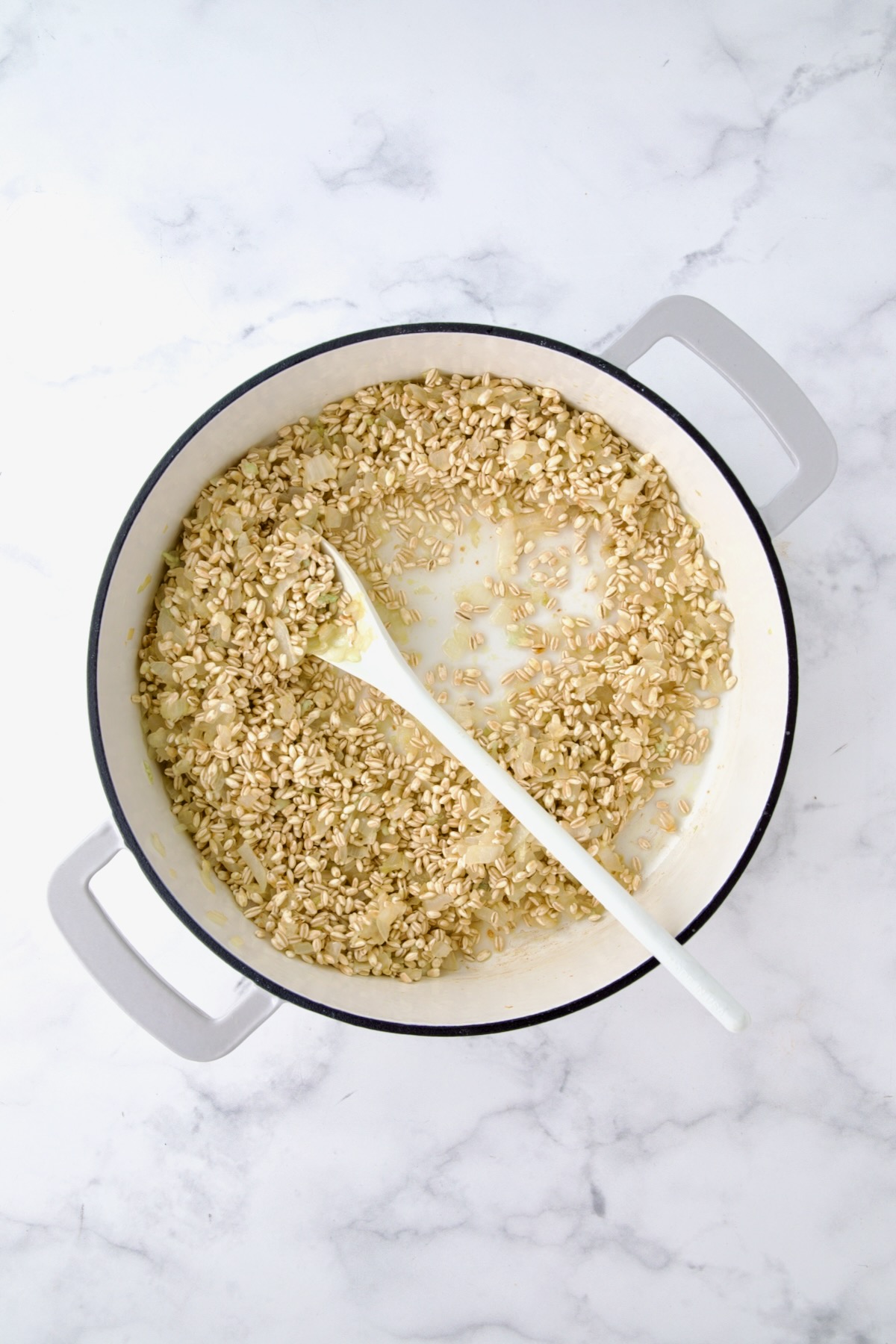 Cooked chopped onions, garlic and uncooked barley in white enamel cast iron skillet after wine is added being stirred with white mixing spoon.