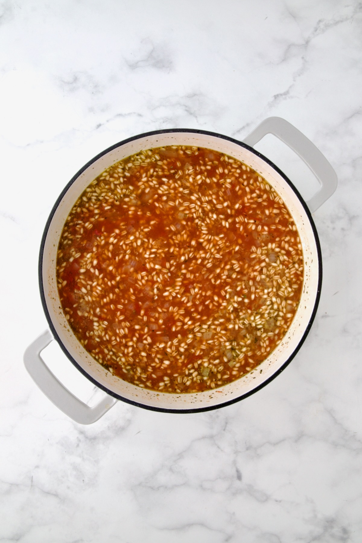 Vegetable broth, crushed tomatoes and Italian seasoning added to barley, onions and garlic stirred together in white enamel cast iron skillet.