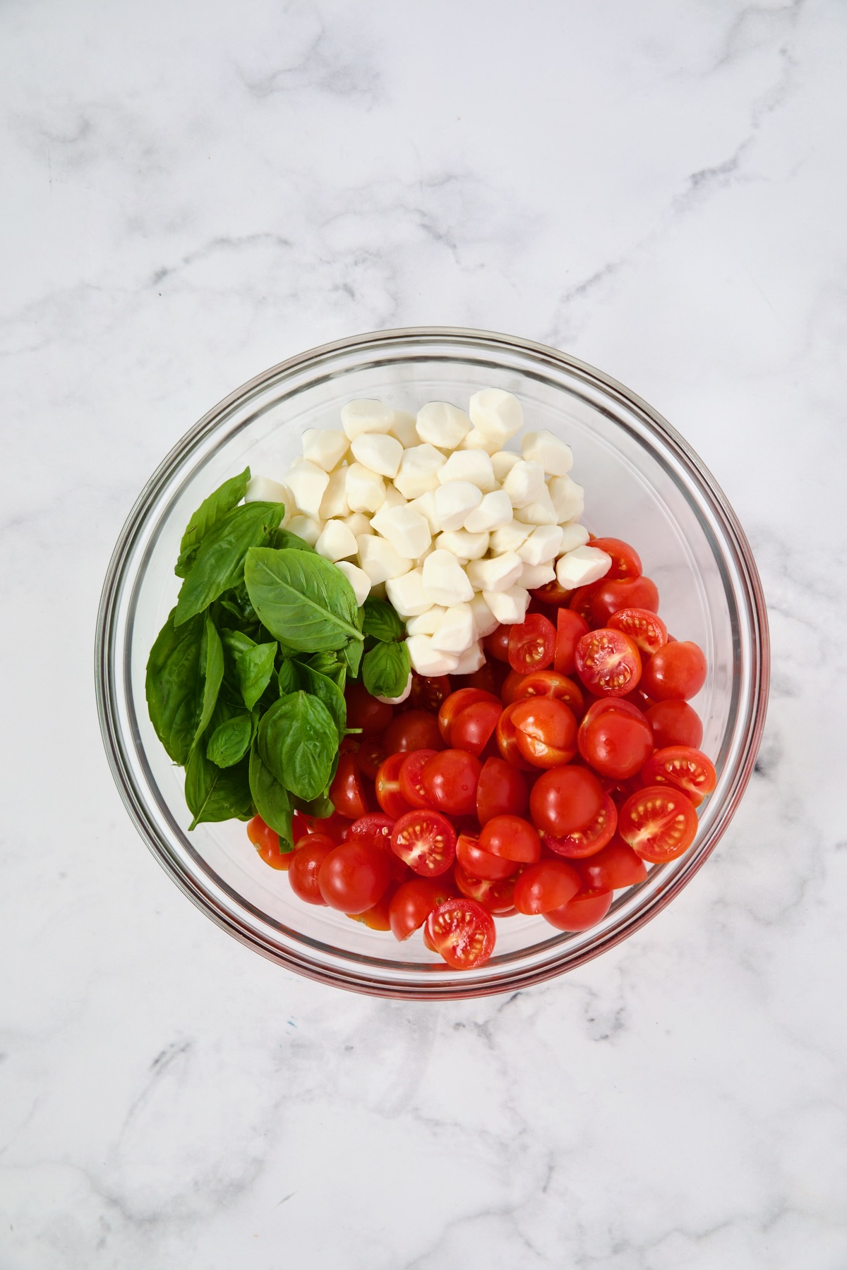 Fresh basil, mozzarella pearls and halved cherry tomatoes in glass bowl before being stirred together.