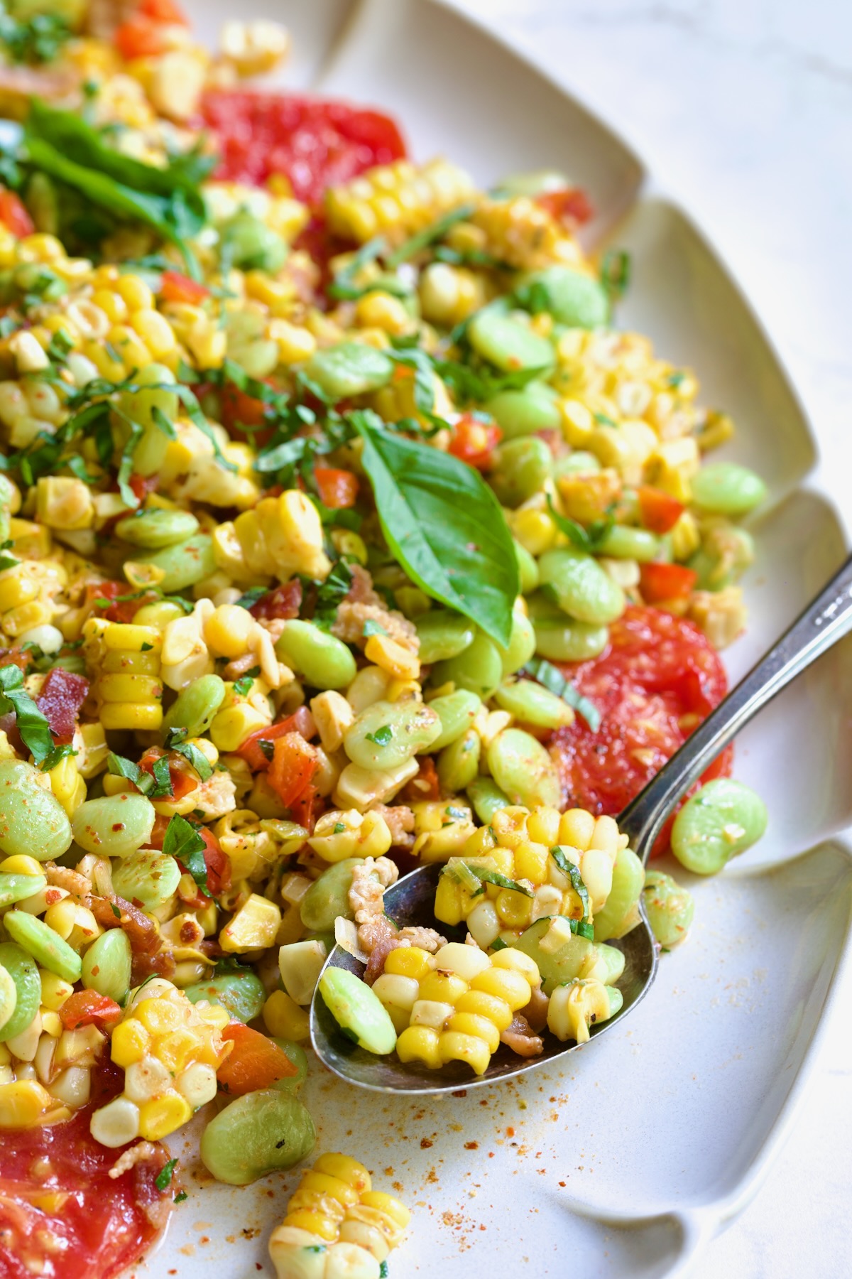 Grilled Corn Succotash garnished with fresh basil on white oval platter with serving spoon scooping some up.