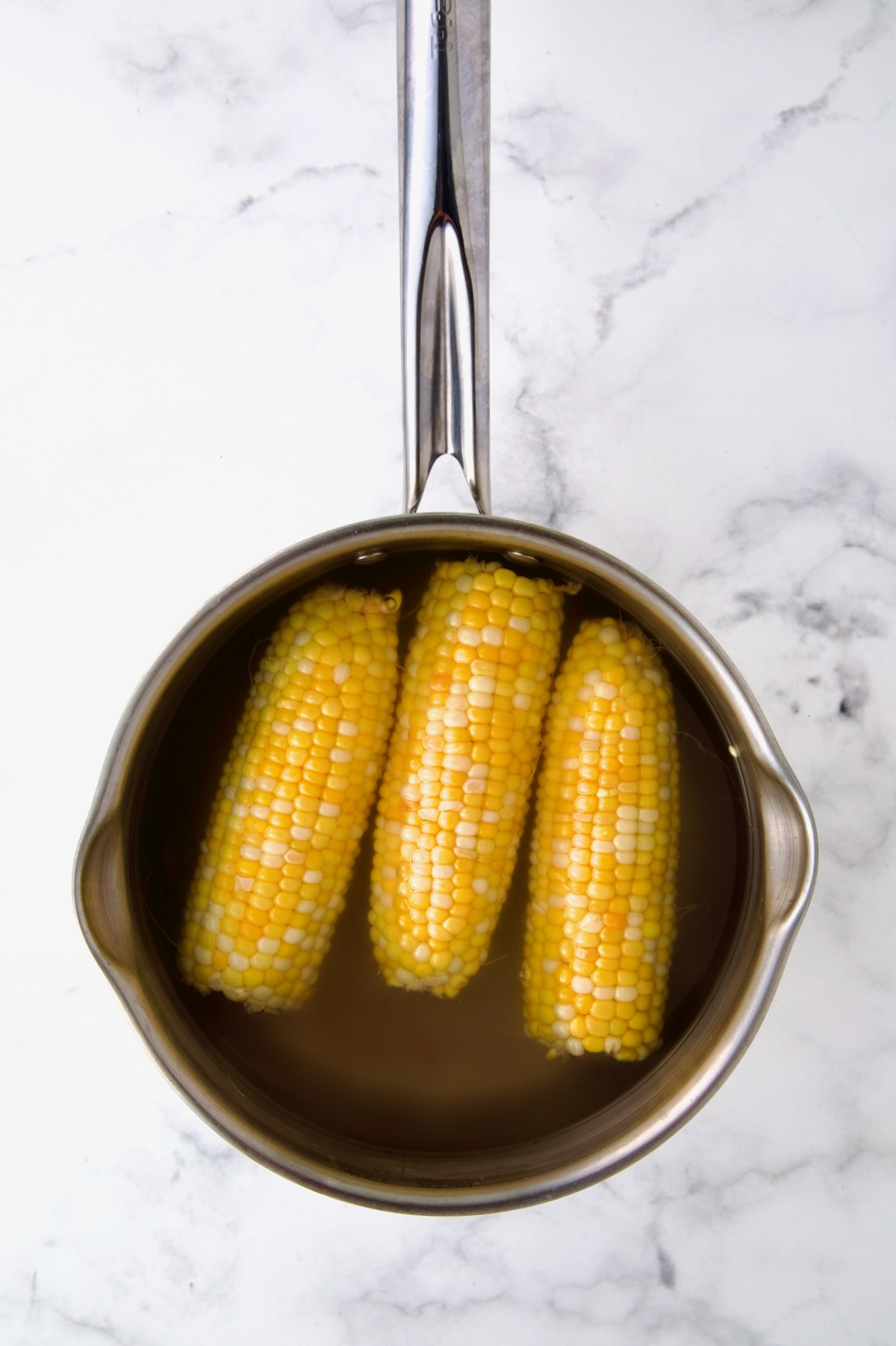 Three ears of corn in boiling water in stainless steel saucepan.