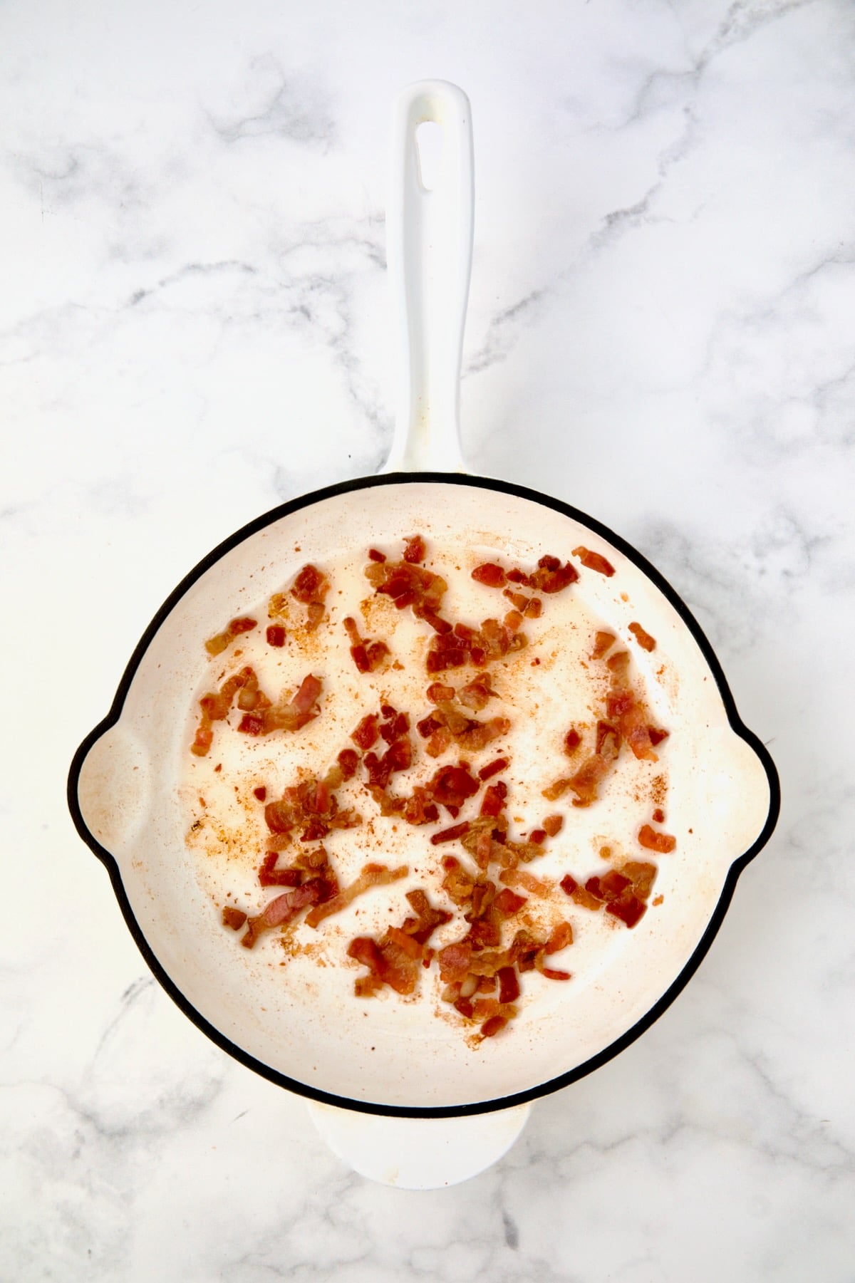 Bacon being crisped in white enamel cast iron skillet.
