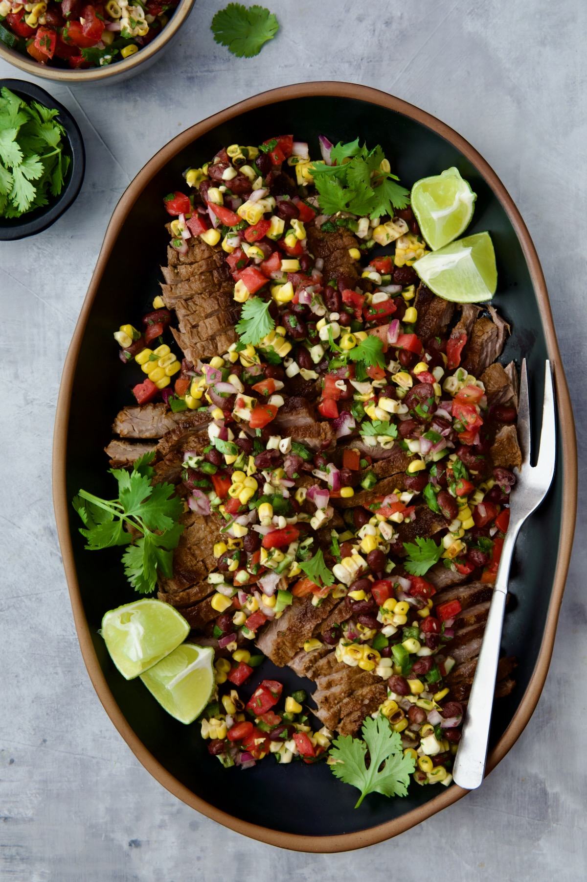 Grilled Skirt Steak with Black Bean Corn Tomato Salad on black platter garnished with lime wedges and cilantro sprigs.