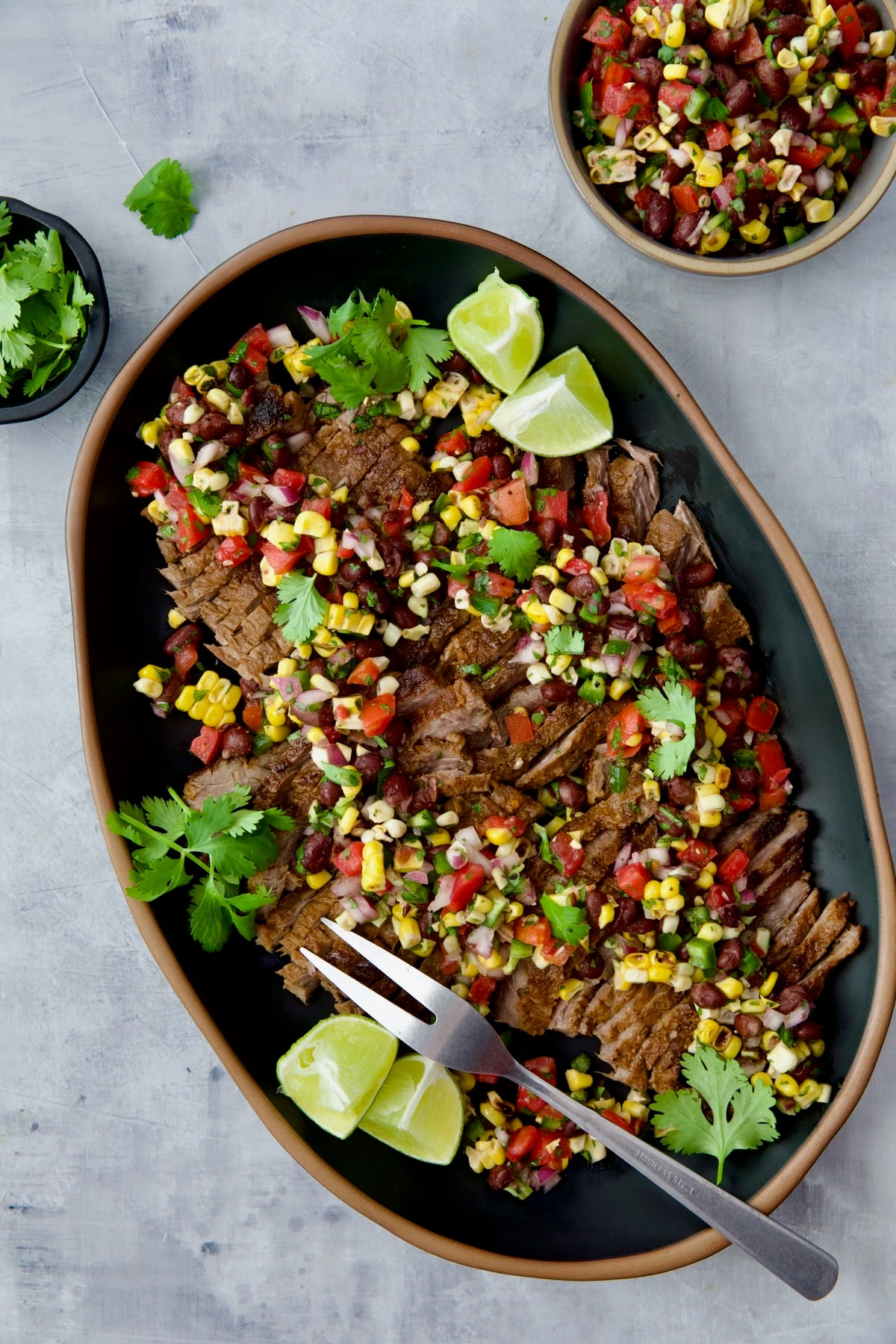 Grilled Skirt Steak with Black Bean Corn Tomato Salad on black platter garnished with lime wedges and cilantro sprigs.