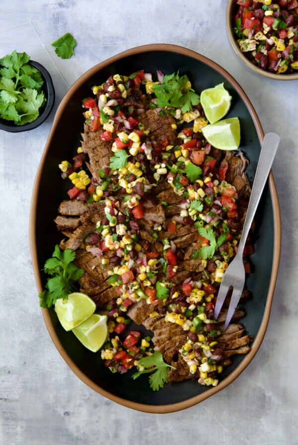 Grilled Skirt Steak with Black Bean Corn Tomato Salad on black platter garnished with lime wedges and cilantro sprigs.