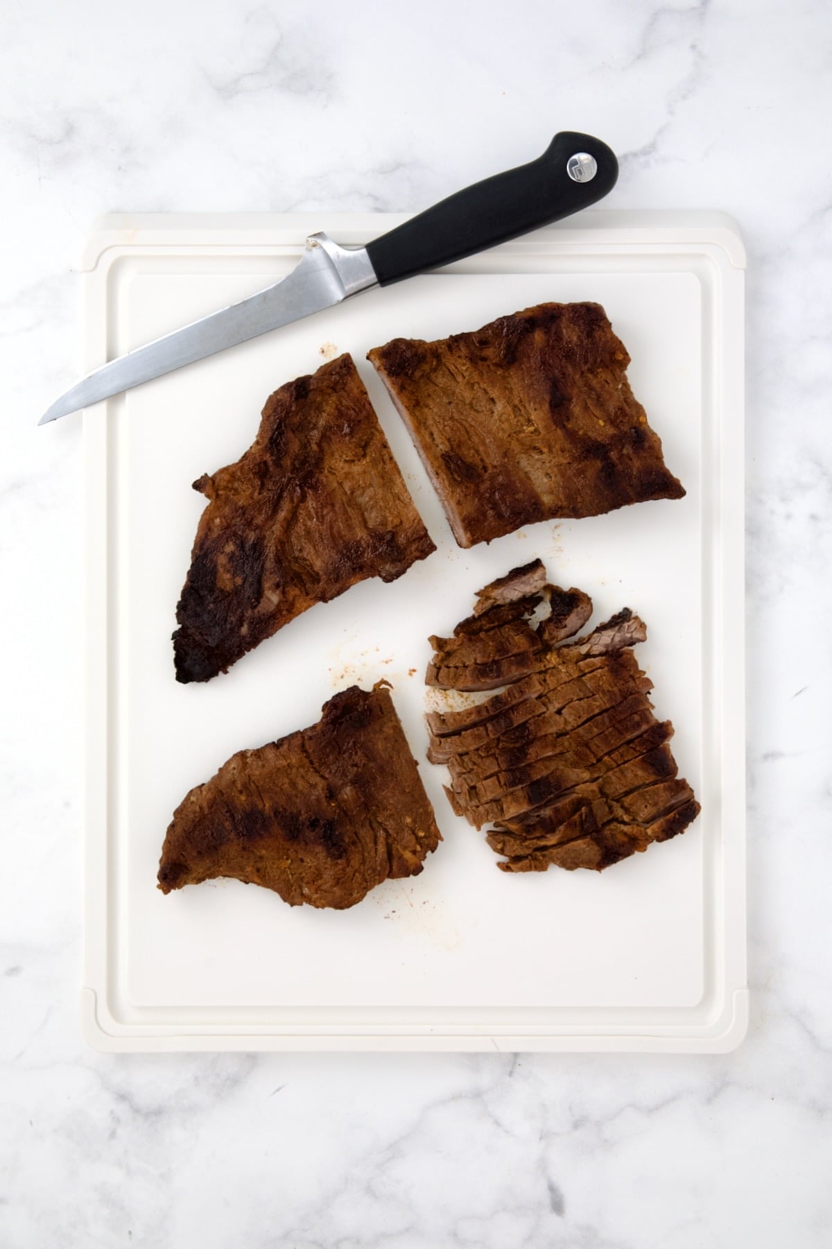 Two pieces of grilled skirt steak on white plastic cutting board being sliced.