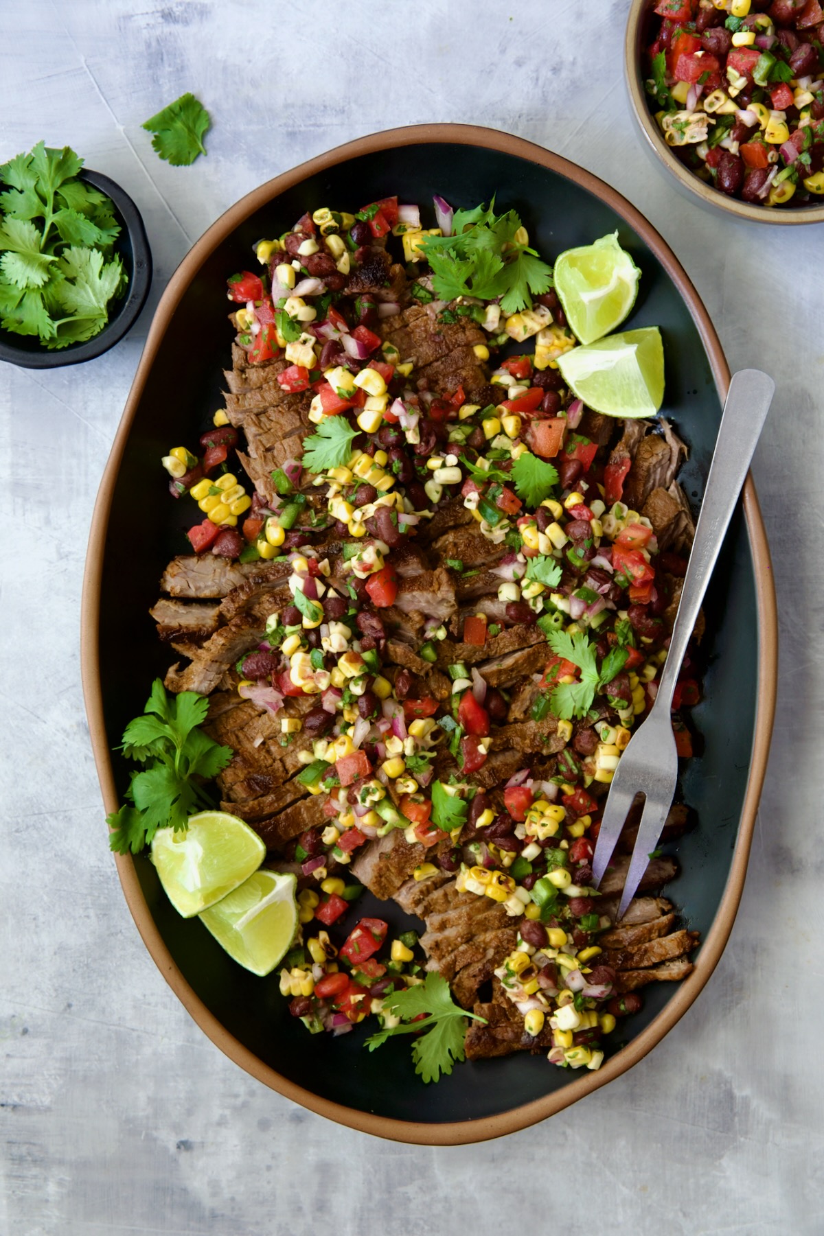 Grilled Skirt Steak with Black Bean Corn Tomato Salad on black platter garnished with lime wedges and cilantro sprigs.