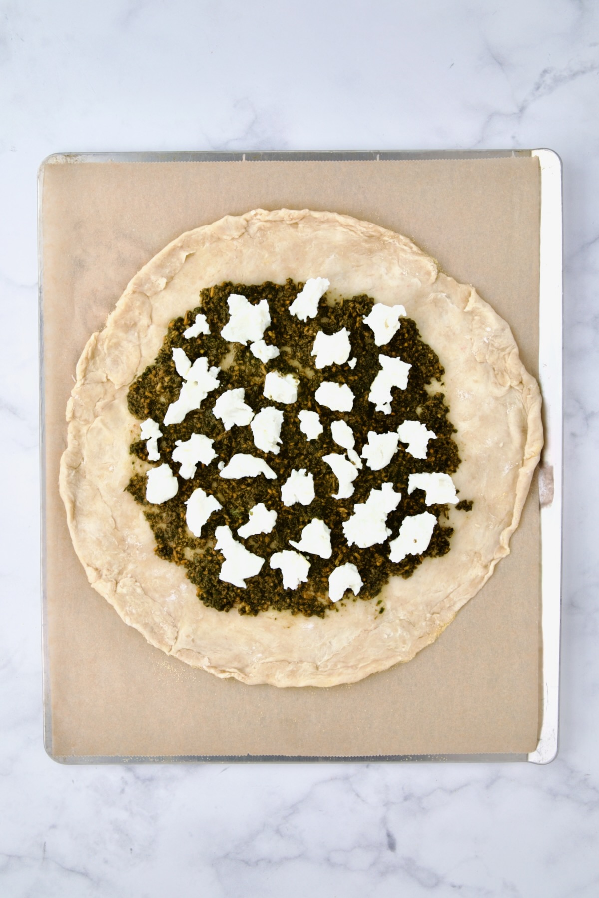 Rolled out galette dough on parchment paper on cookie sheet with pesto and goat cheese added.