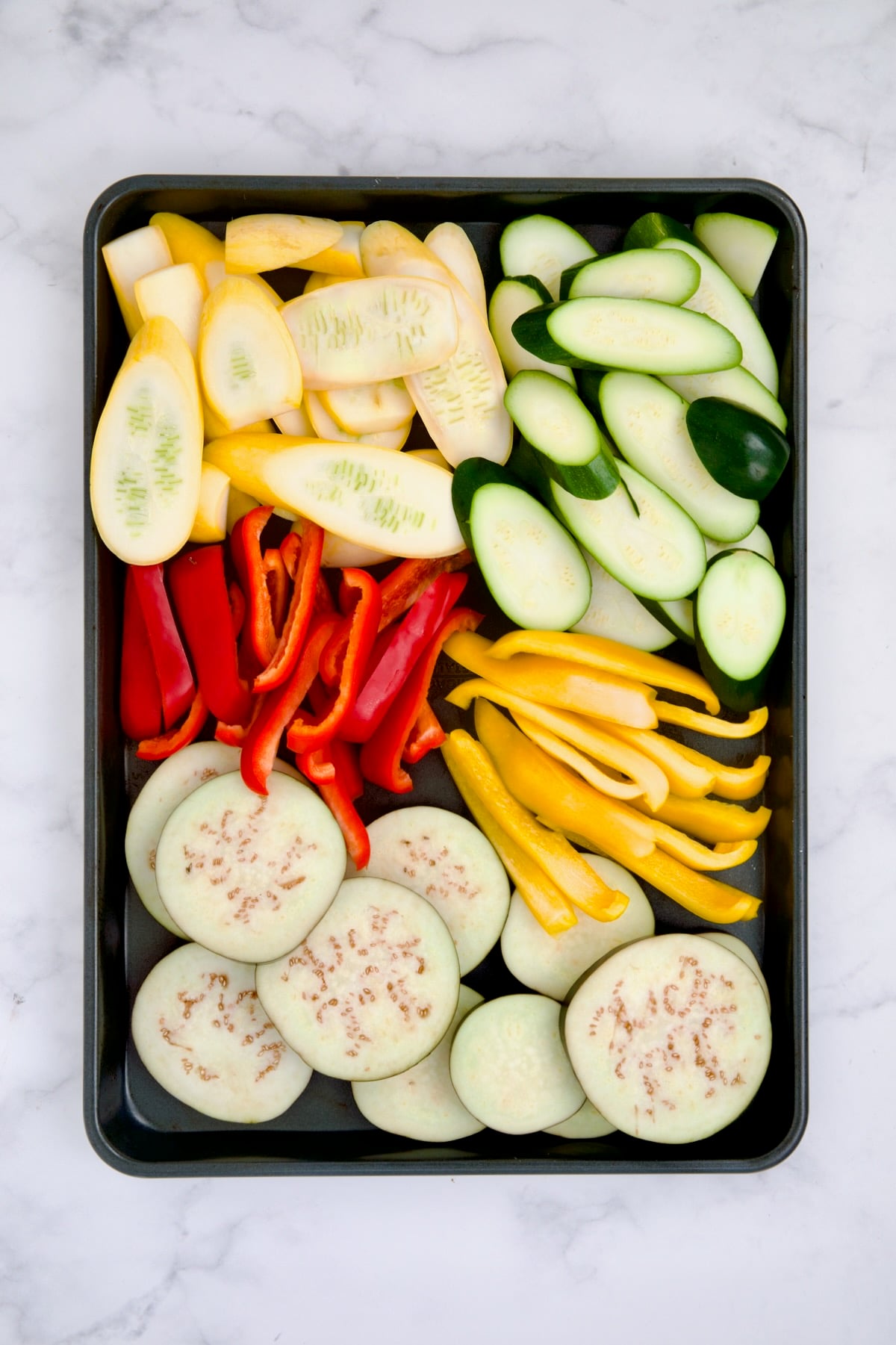Yellow squash, zucchini, red bell pepper, yellow bell pepper and eggplant on rimmed sheet pan before being grilled.