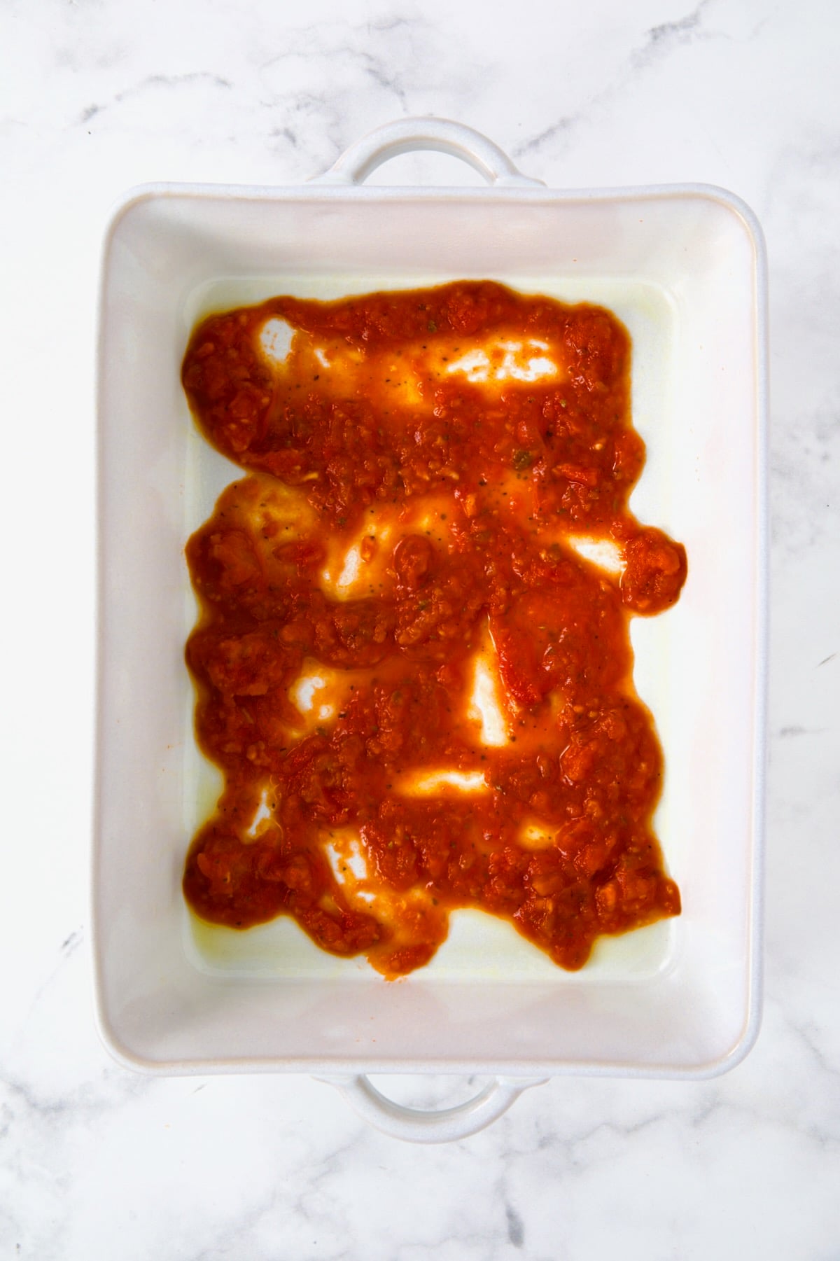 Layer of marinara sauce in white rectangular baking dish.
