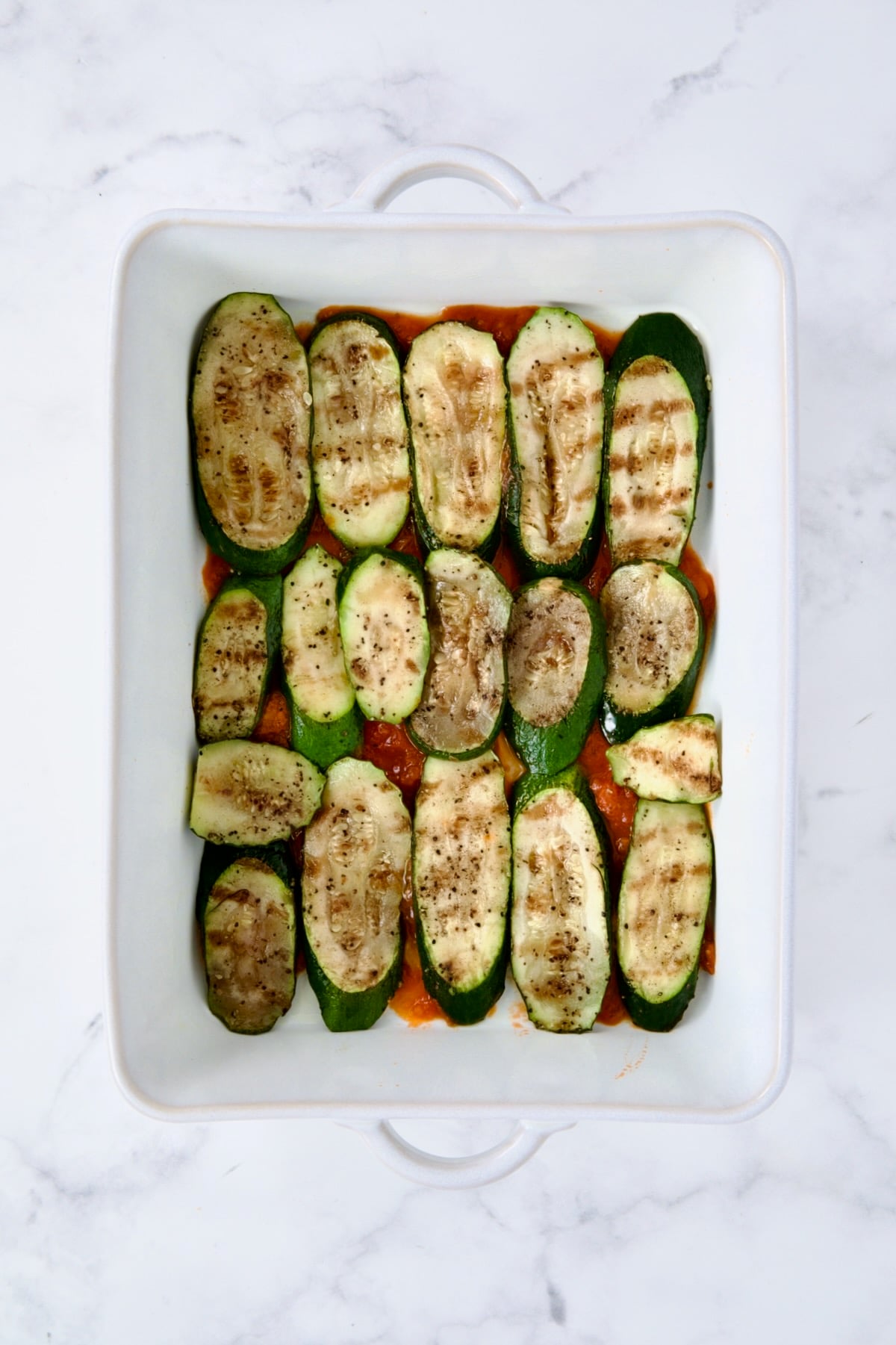 Layer of zucchini over marinara sauce in white rectangular baking dish.