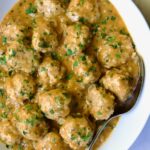 Baked Turkey Meatballs in Beer - Onion Sauce in white oval bowl with serving spoon garnished with fresh parsley.