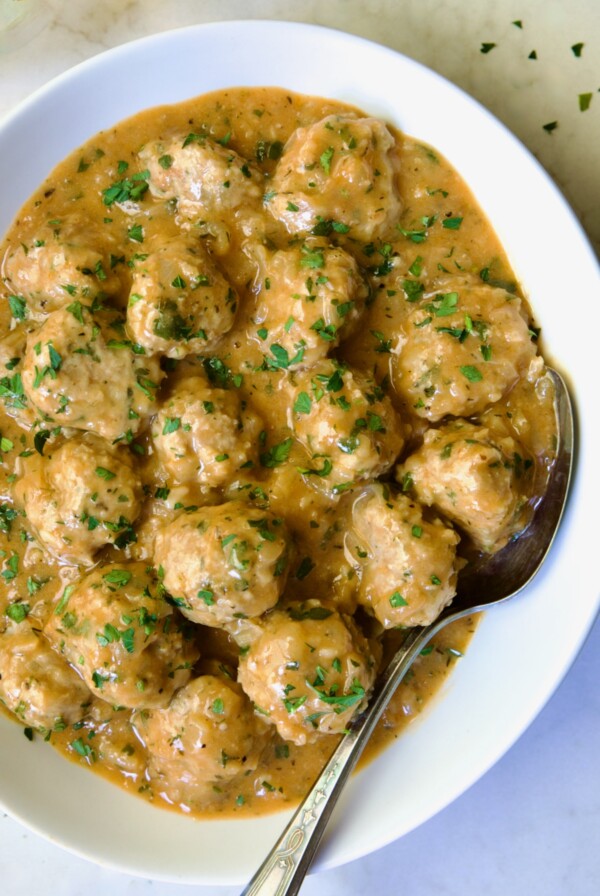 Baked Turkey Meatballs in Beer - Onion Sauce in white oval bowl with serving spoon garnished with fresh parsley.