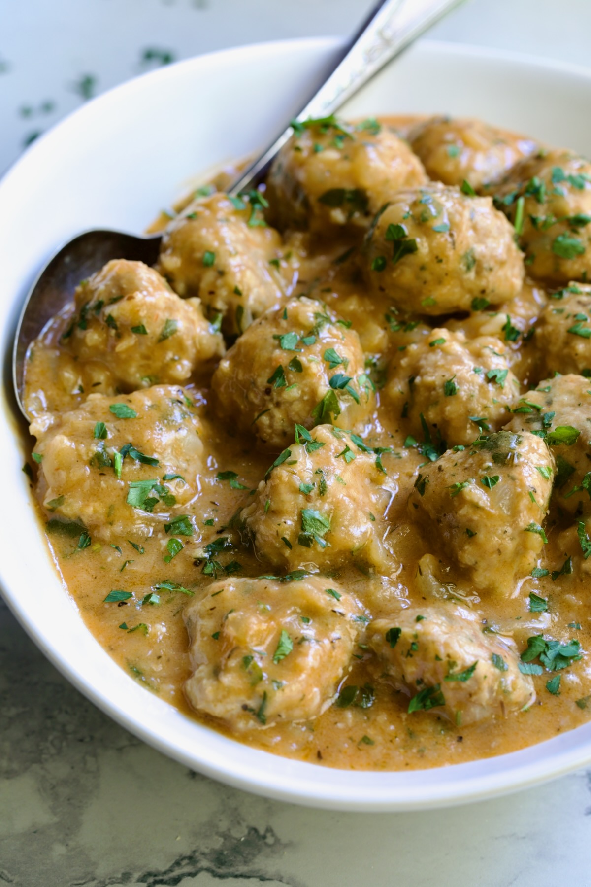 Baked Turkey Meatballs in Beer - Onion Sauce in white oval bowl with serving spoon garnished with fresh parsley.