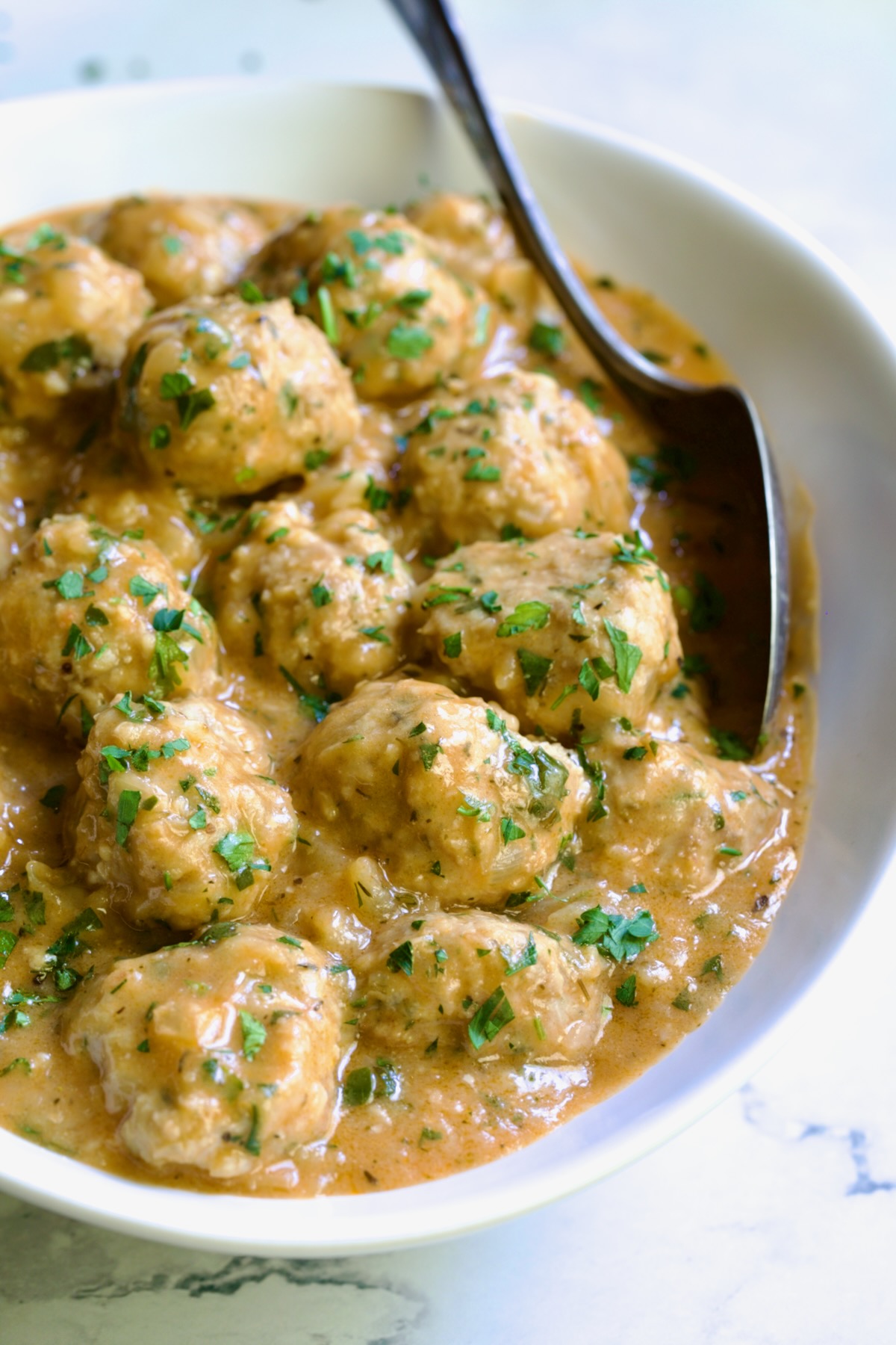 Baked Turkey Meatballs in Beer - Onion Sauce in white oval bowl with serving spoon garnished with fresh parsley.