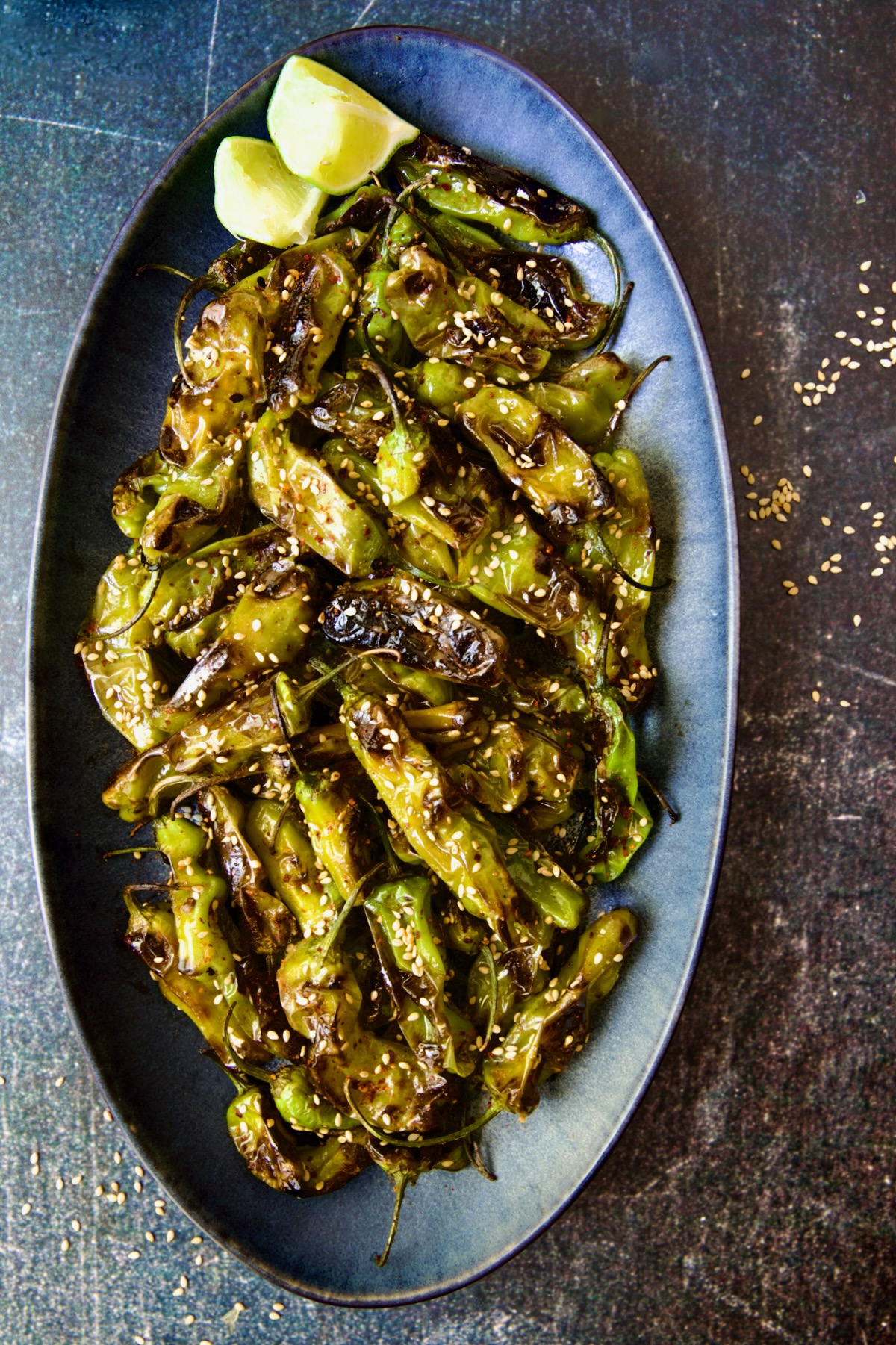 Blistered Shishito Peppers on blue oval platter garnished with sesame seeds and lime wedges.