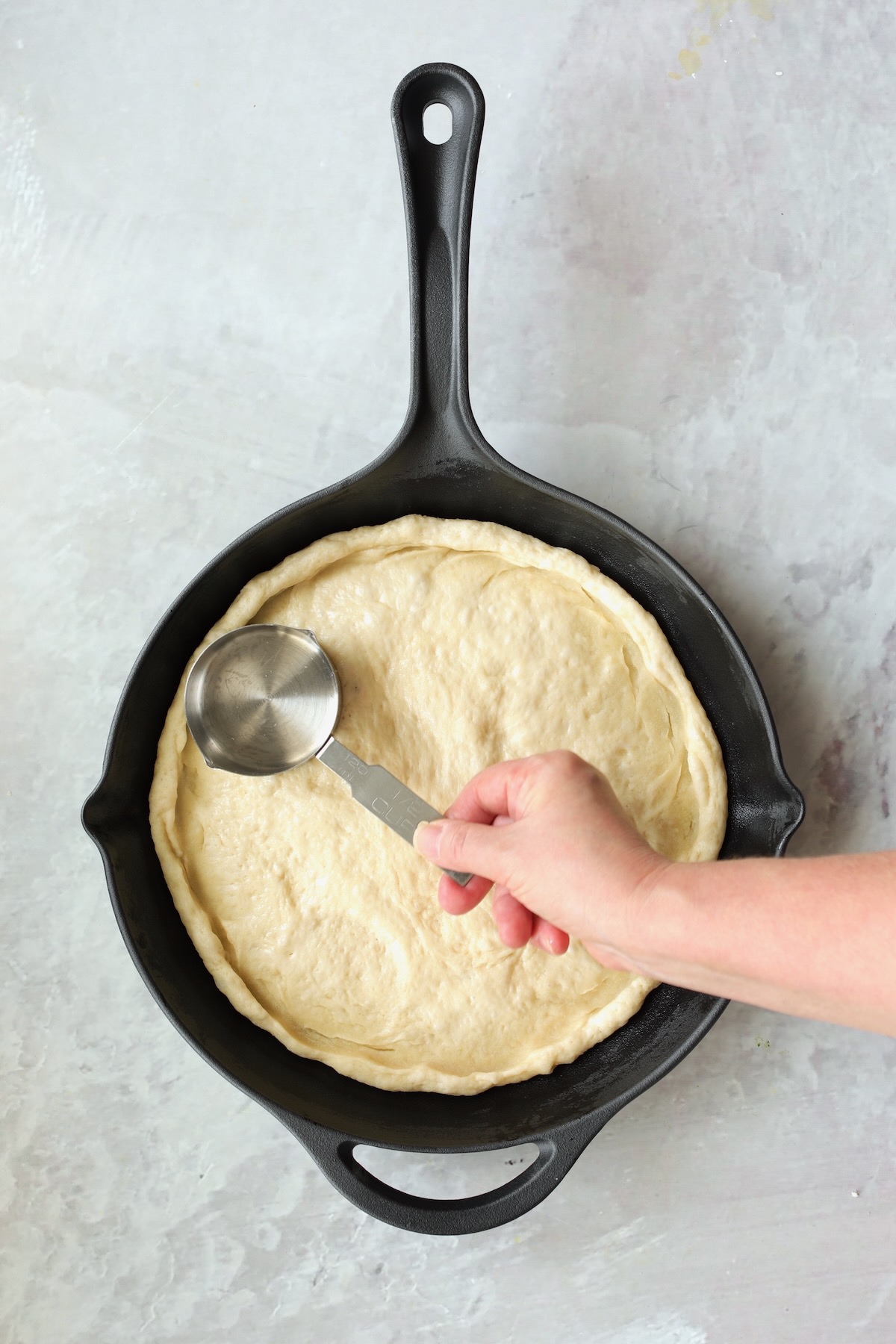 Pizza dough in cast iron skillet being formed to the skillet with 1-cup measuring cup.