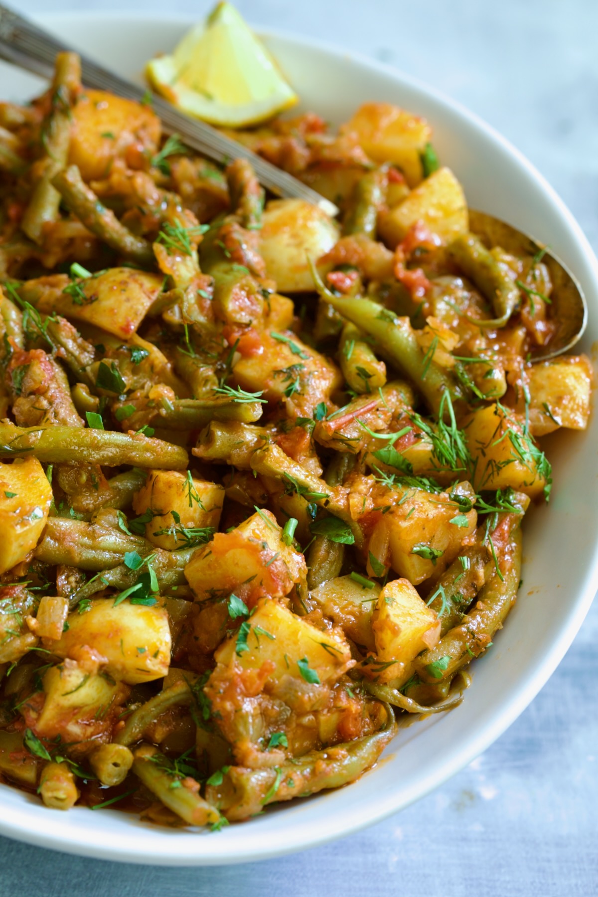 Fasolakia in oval white serving bowl garnished with fresh herbs and lemon wedges.