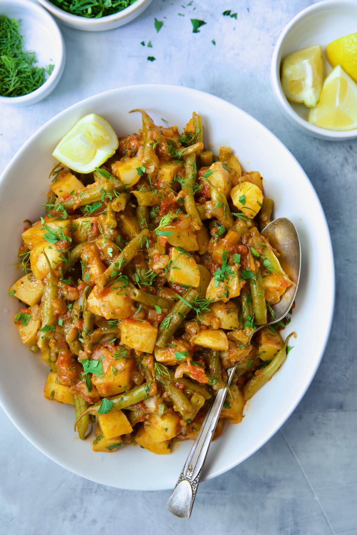 Fasolakia in oval white serving bowl garnished with fresh herbs and lemon wedges.