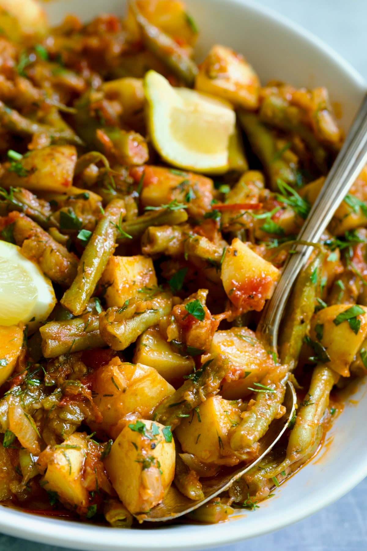 Fasolakia in oval white serving bowl garnished with fresh herbs and lemon wedges.
