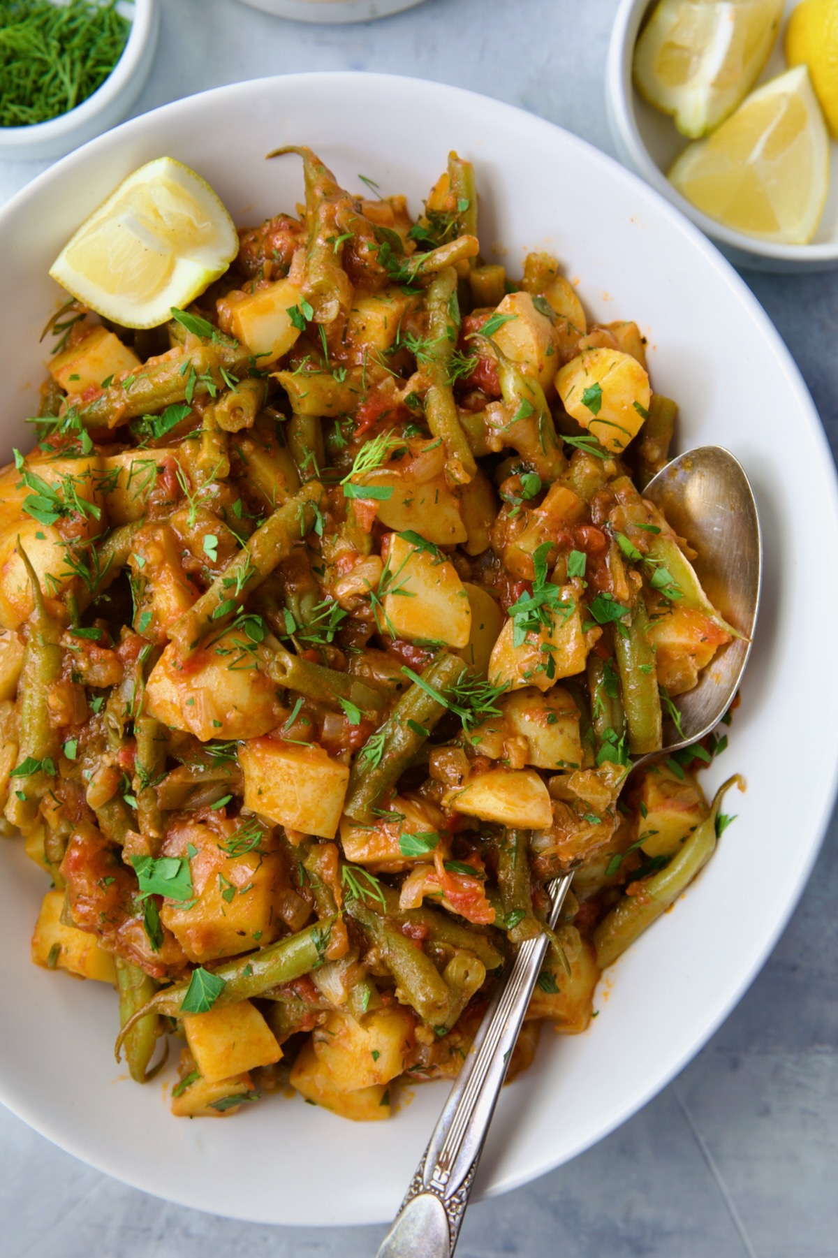 Fasolakia in oval white serving bowl garnished with fresh herbs and lemon wedges.