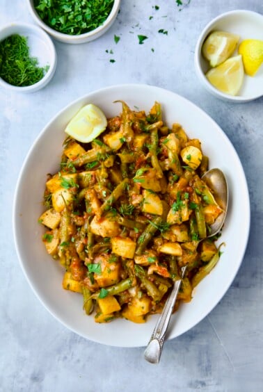 Fasolakia in oval white serving bowl garnished with fresh herbs and lemon wedges.