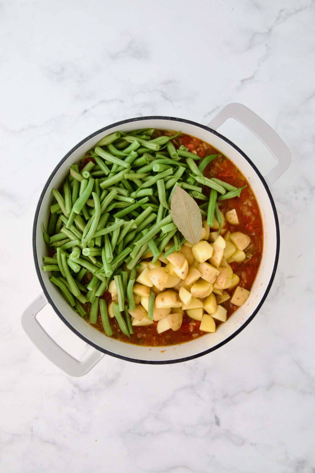 Potatoes, green beans and bay leaf added to tomato sauce in white enamel cast iron skillet.