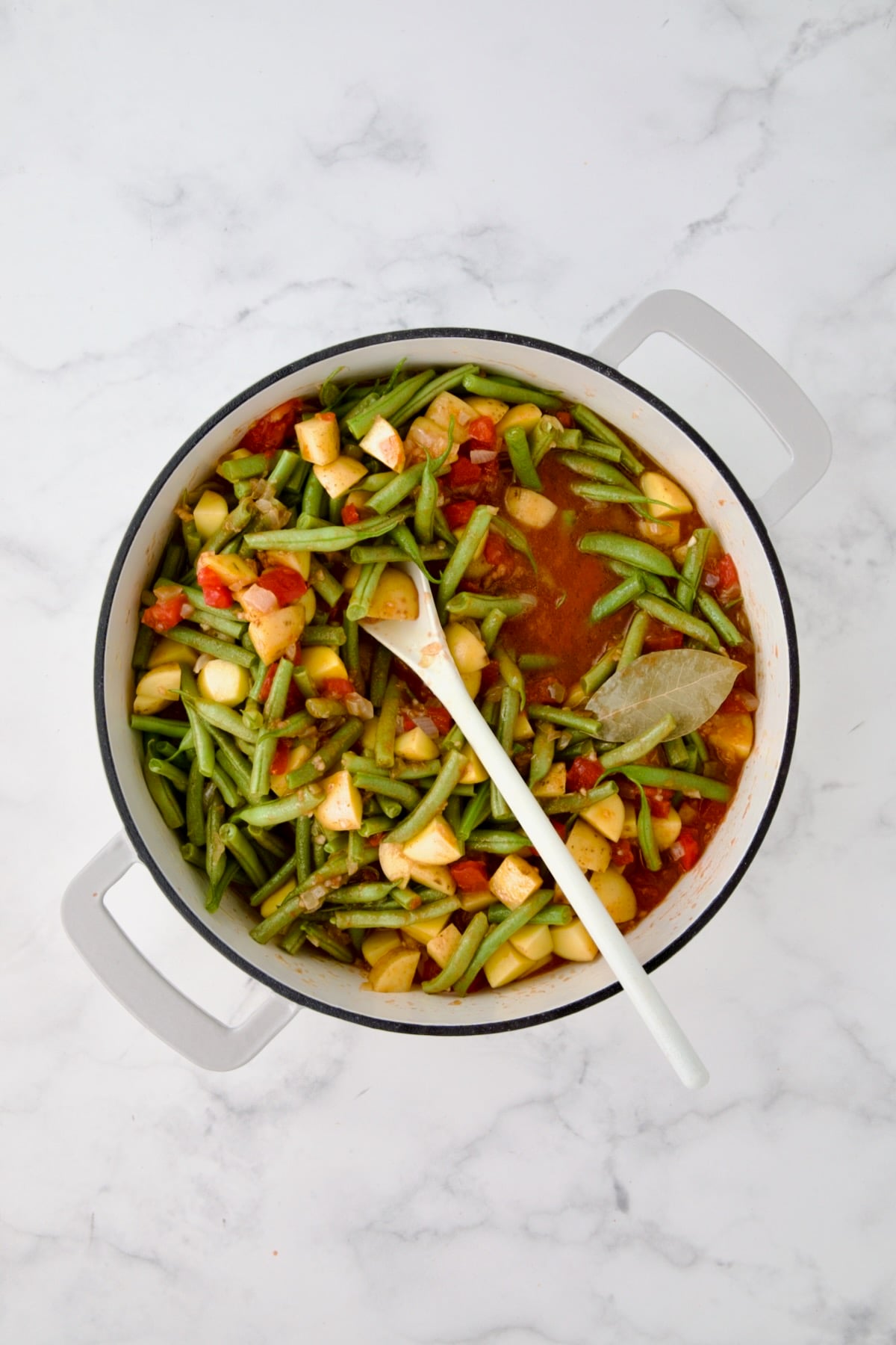 Potatoes, green beans and bay leaf added to tomato sauce being mixed together with white spoon in white enamel cast iron skillet.