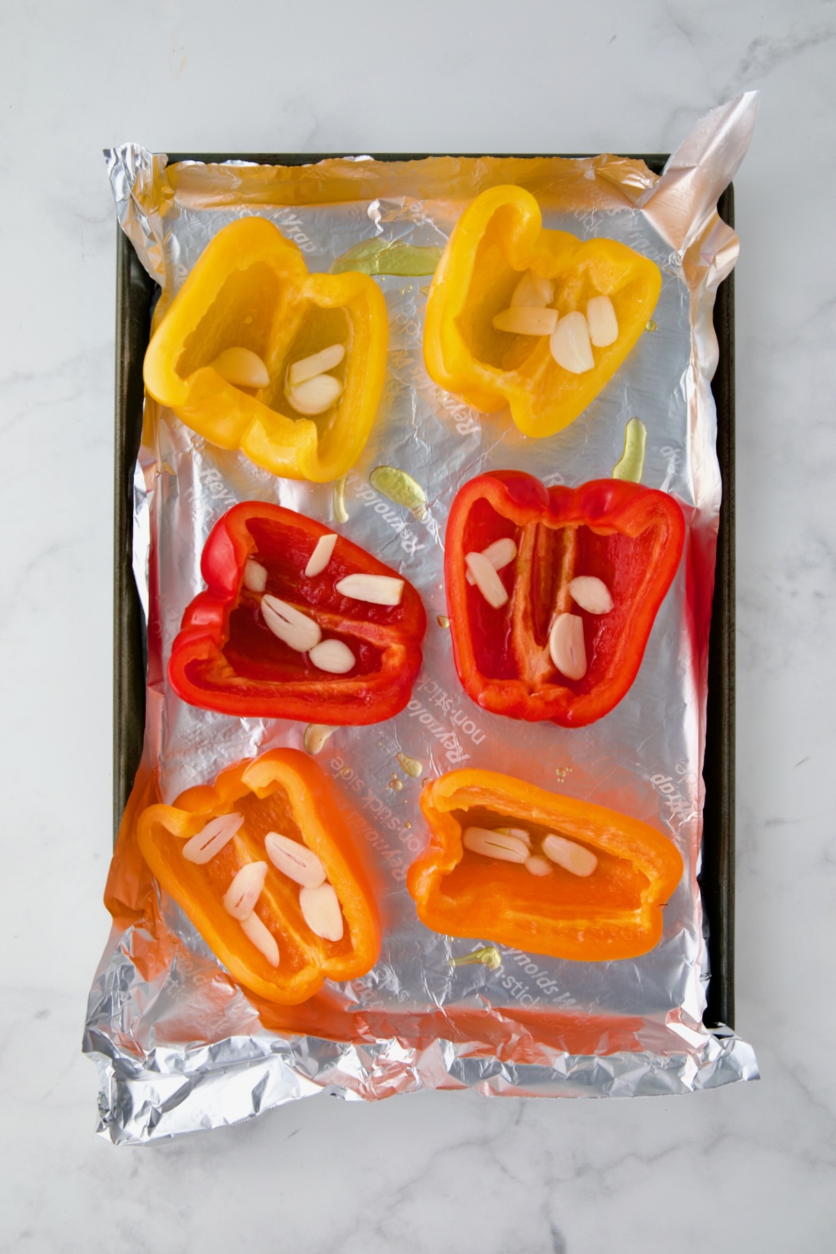 Yellow, red and orange bell pepper halves on aluminum foil-lined rimmed sheet pan with garlic halves in them.