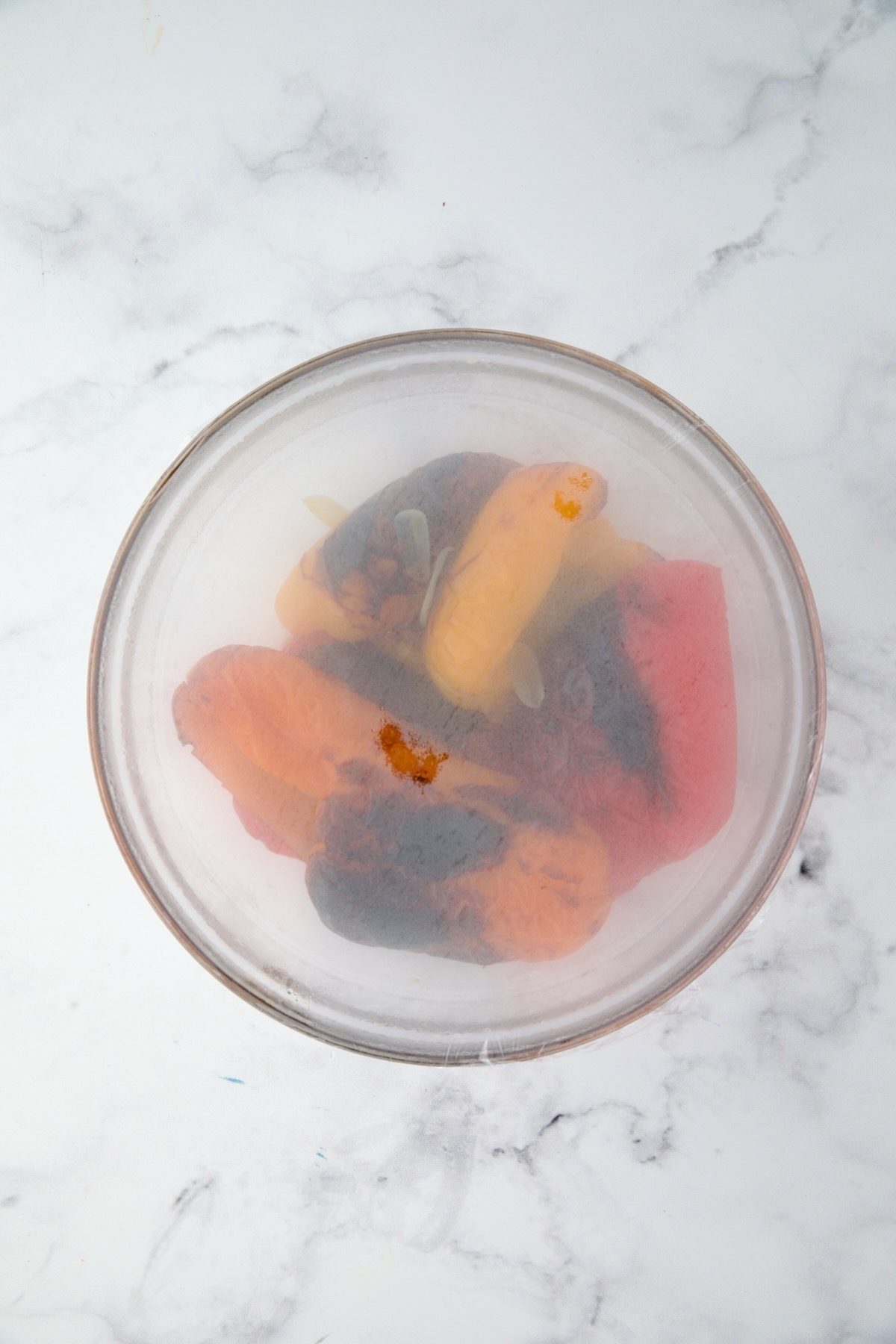 Roasted bell pepper halves in glass bowl with plastic wrap over it for the peppers to steam.
