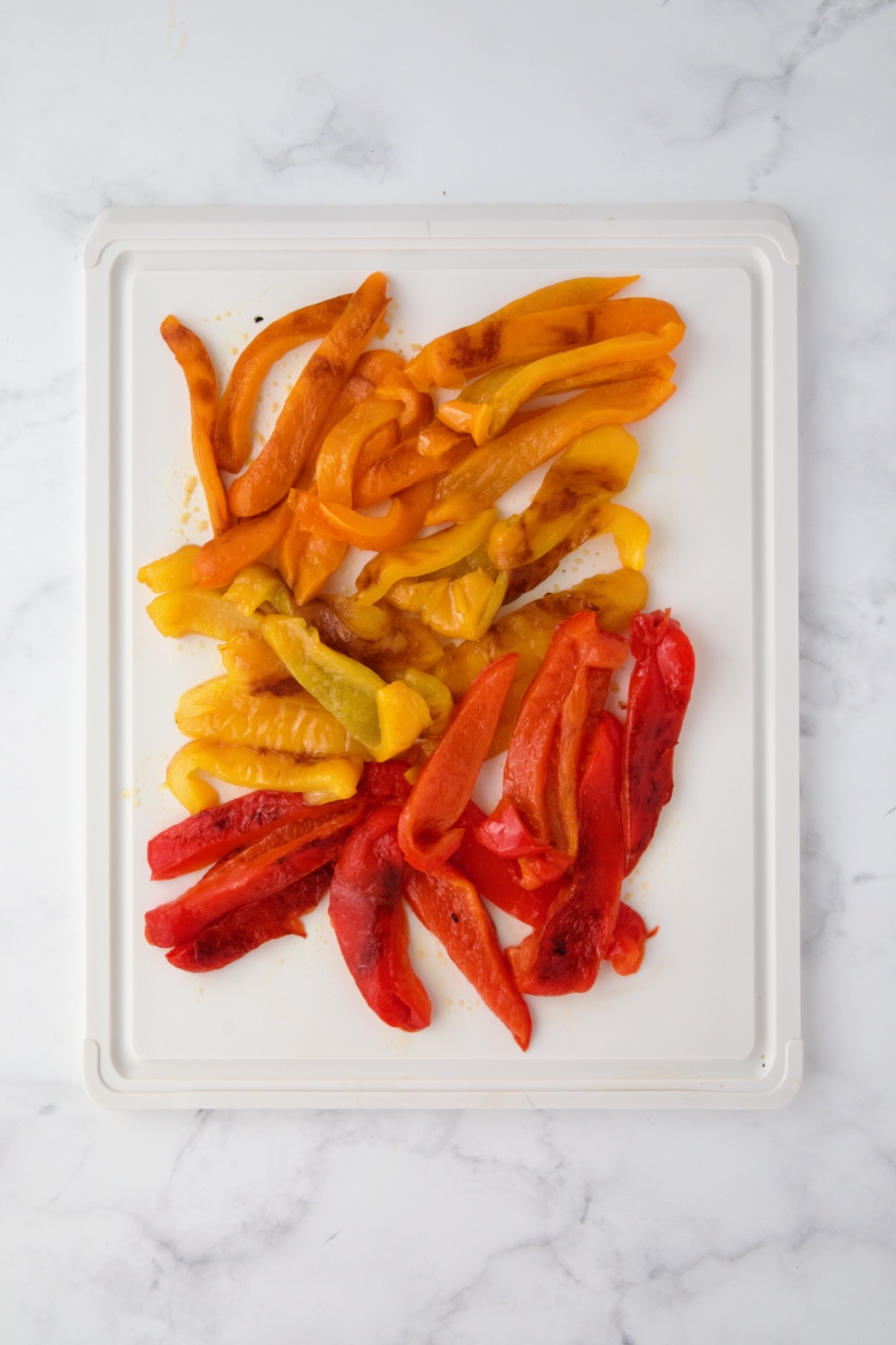 Orange, yellow and red bell pepper halves cut into strips on white plastic cutting board.