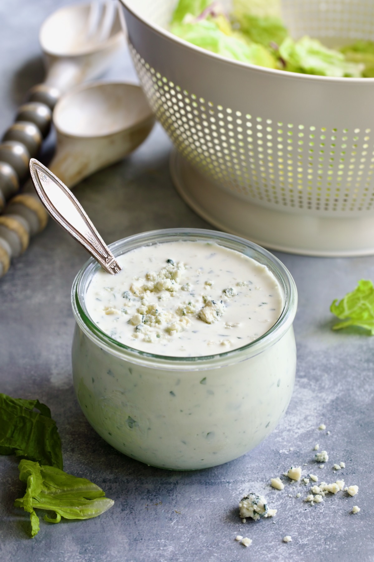 Best Blue Cheese Dressing Recipe in glass jar with spoon and colander of lettuce in the background.